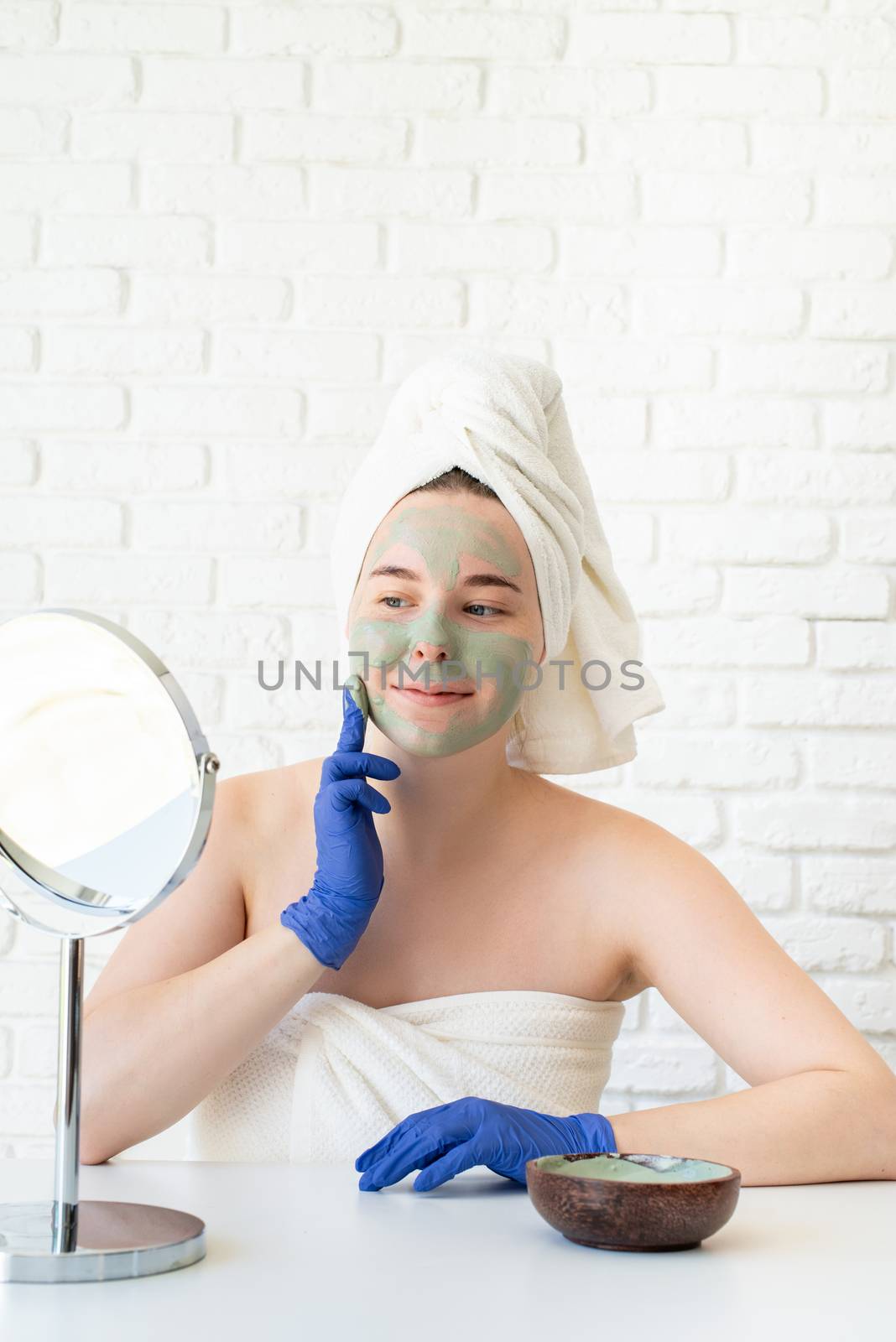 Spa and wellness. Natural cosmetics. Self care. Close up of happy young caucasian woman in white bath towels wearing gloves applying clay face mask looking at the mirror