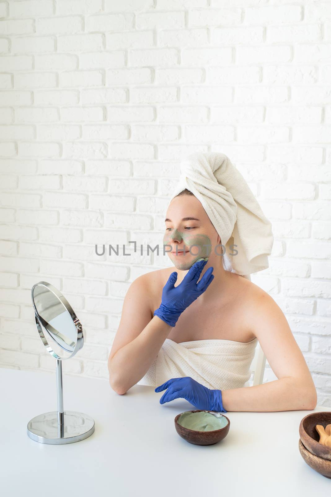 Spa and wellness. Natural cosmetics. Self care. Close up of happy young caucasian woman in white bath towels wearing gloves applying clay face mask looking at the mirror