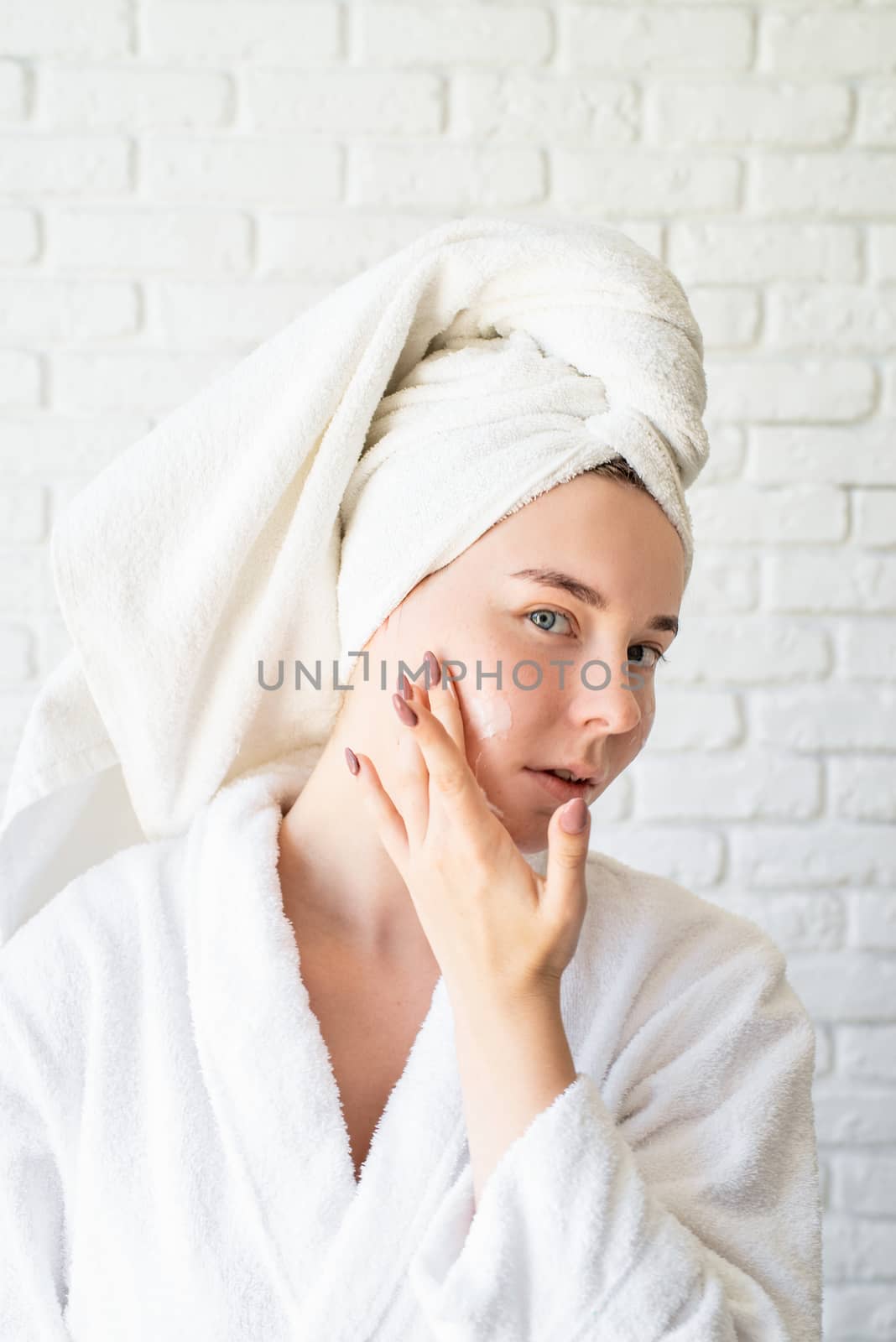 Spa and wellness. Natural cosmetics. Self care. Happy young caucasian woman in white bath towel applying face cream at home