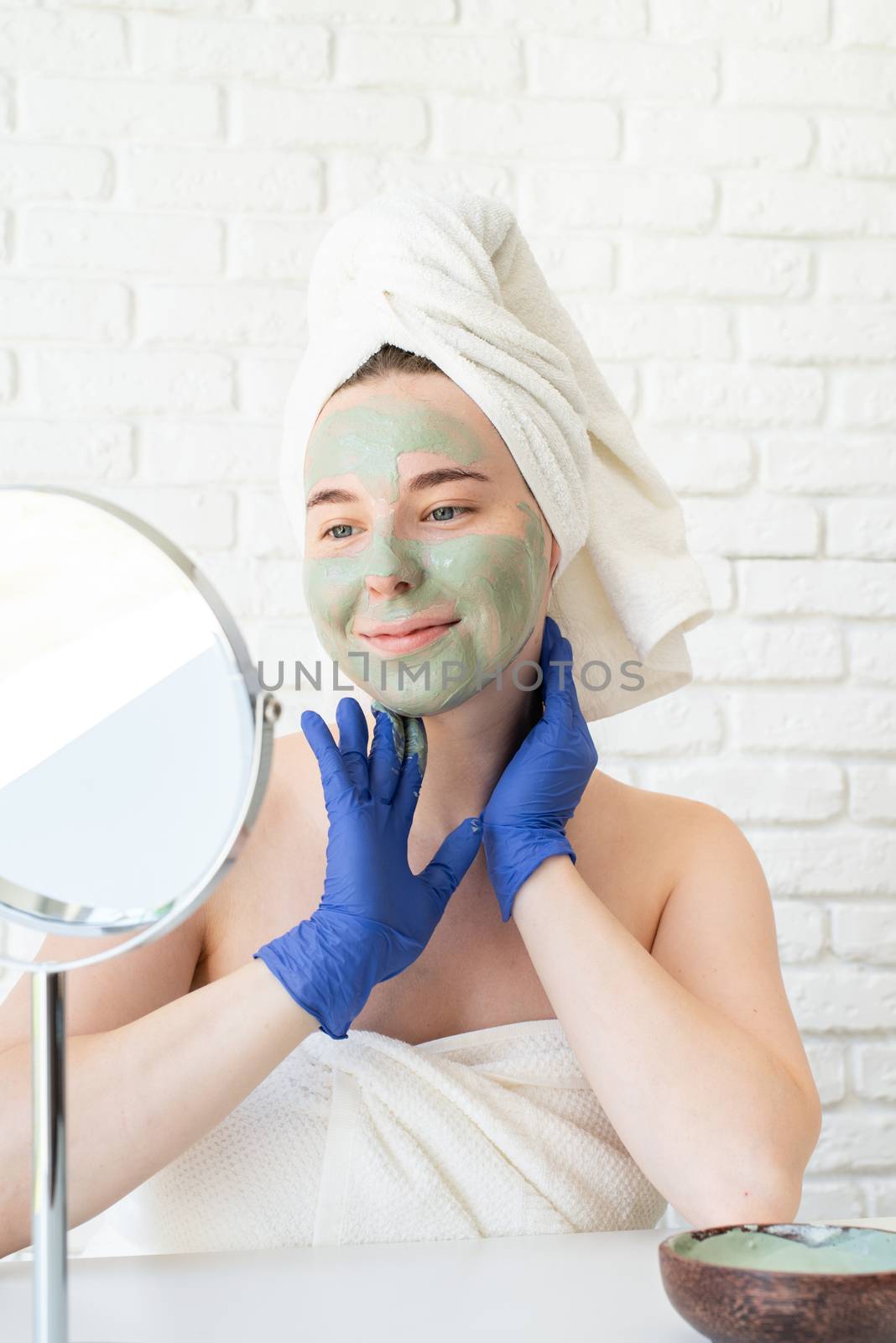 Spa and wellness. Natural cosmetics. Self care. Happy young caucasian woman in white bath towels wearing gloves applying clay face mask looking at the mirror
