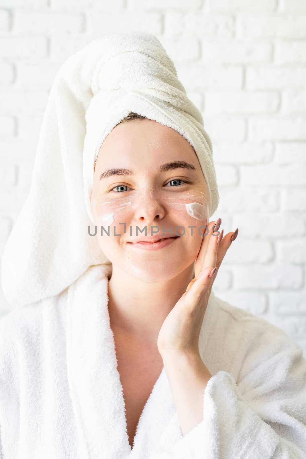Happy young caucasian woman in white bath towel applying face cream at home by Desperada