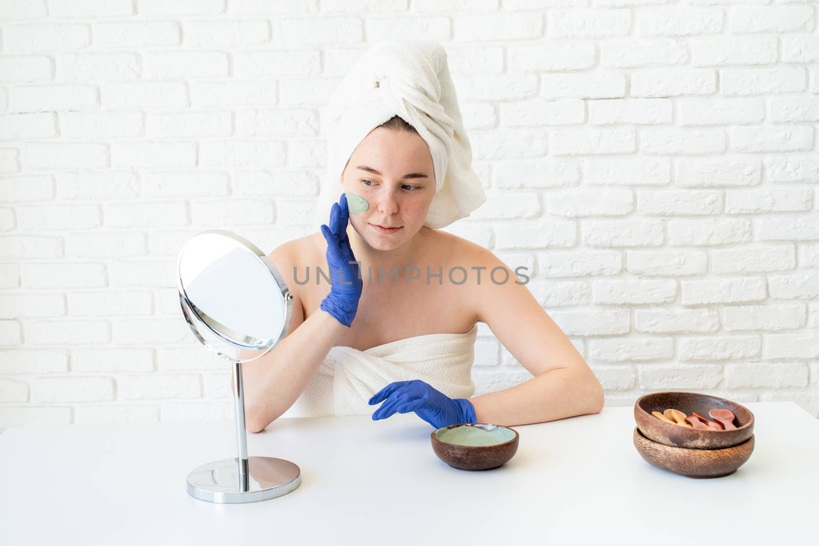 Happy young caucasian woman in white bath towels wearing gloves applying clay face mask looking at the mirror by Desperada