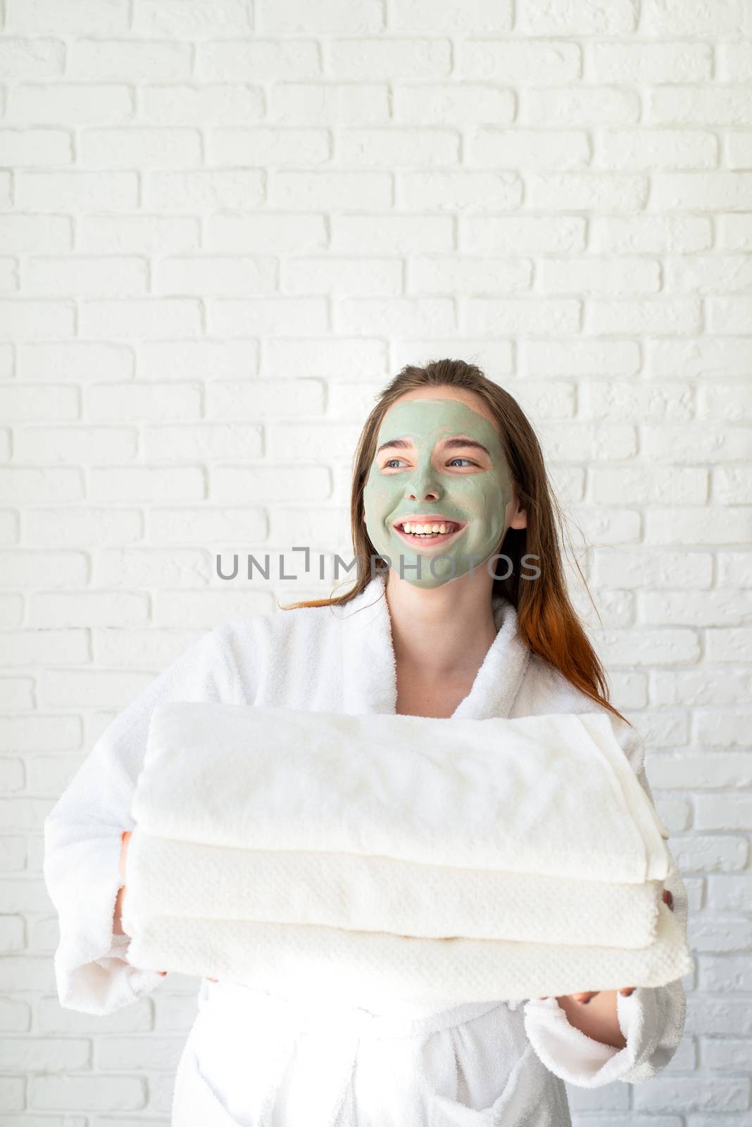 Young smiling caucasian woman with a face mask wearing bathrobes holding a pile of white bath towels by Desperada