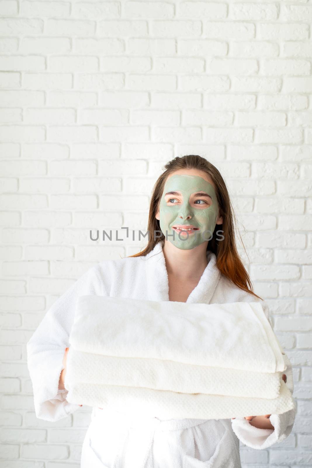 Young smiling caucasian woman with a face mask wearing bathrobes holding a pile of white bath towels by Desperada