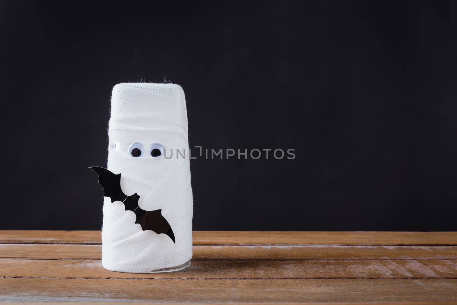 Funny Halloween day decor party concept, The mummy ghost on water glass wrapped around with bandage and have bats and spider stick it found only eyes, studio shot isolated on black background