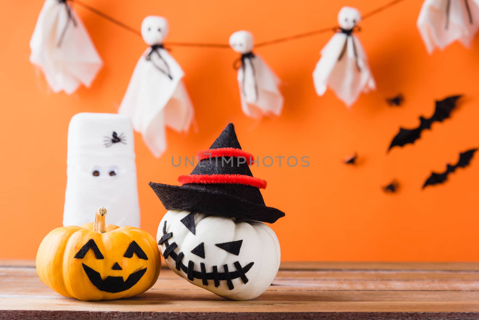 Funny Halloween day decoration party, White ghost scary face, glass of water wrapped with bandage and pumpkin on wood table, studio shot isolated on orange background