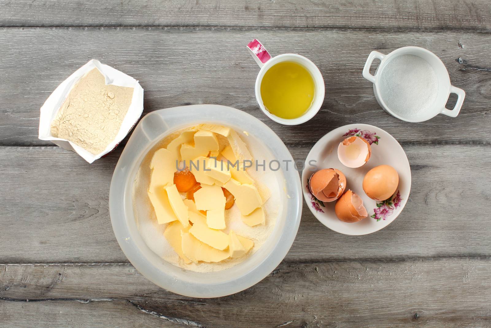 Table top  view on ingredients for cake preparation. Sack of flour, bowl with butter and eggs yolk, cup with glair, egg shells and sugar, placed on gray wood desk.