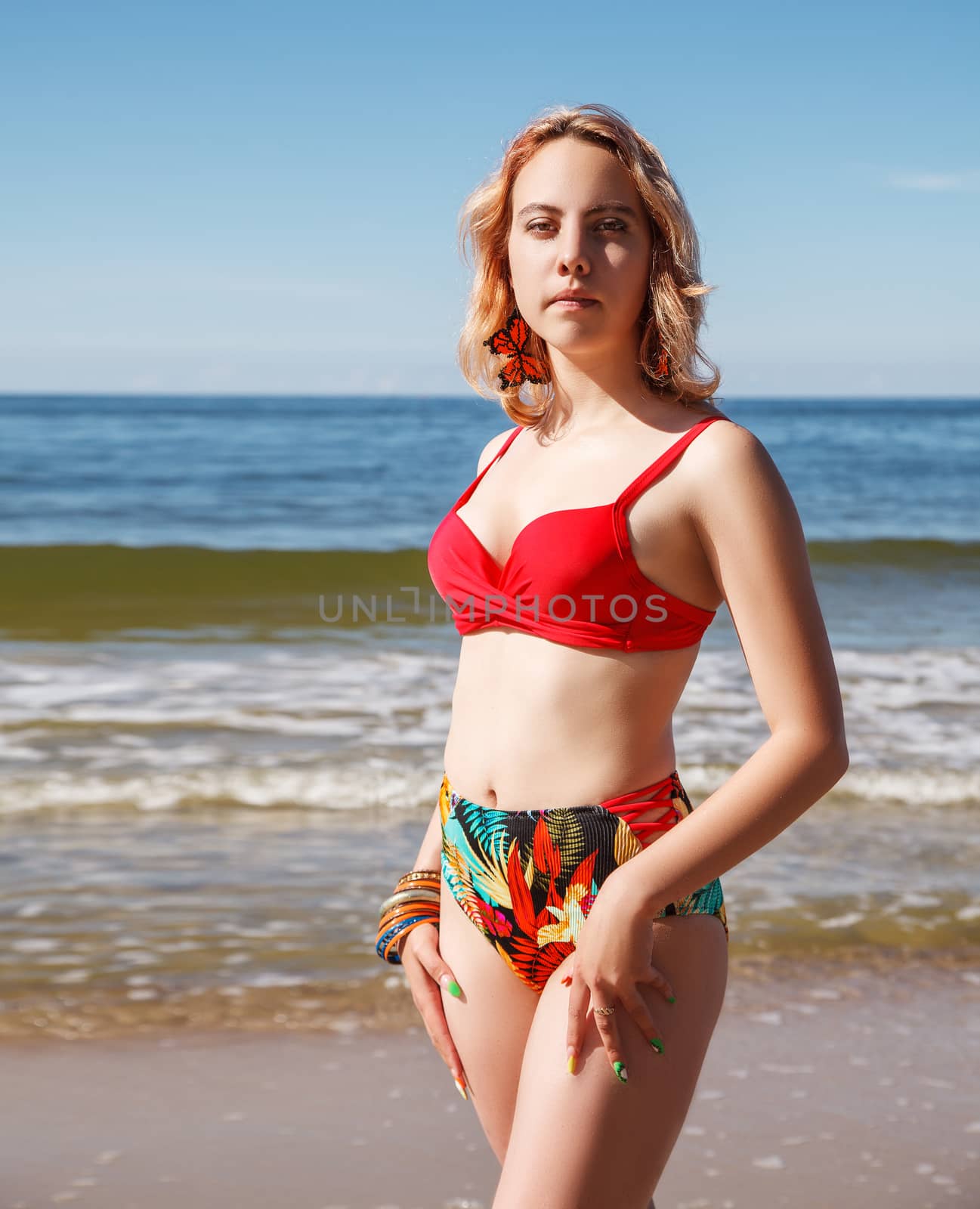 young blonde girl in a red swimsuit standing on the seashore on sunny summer day