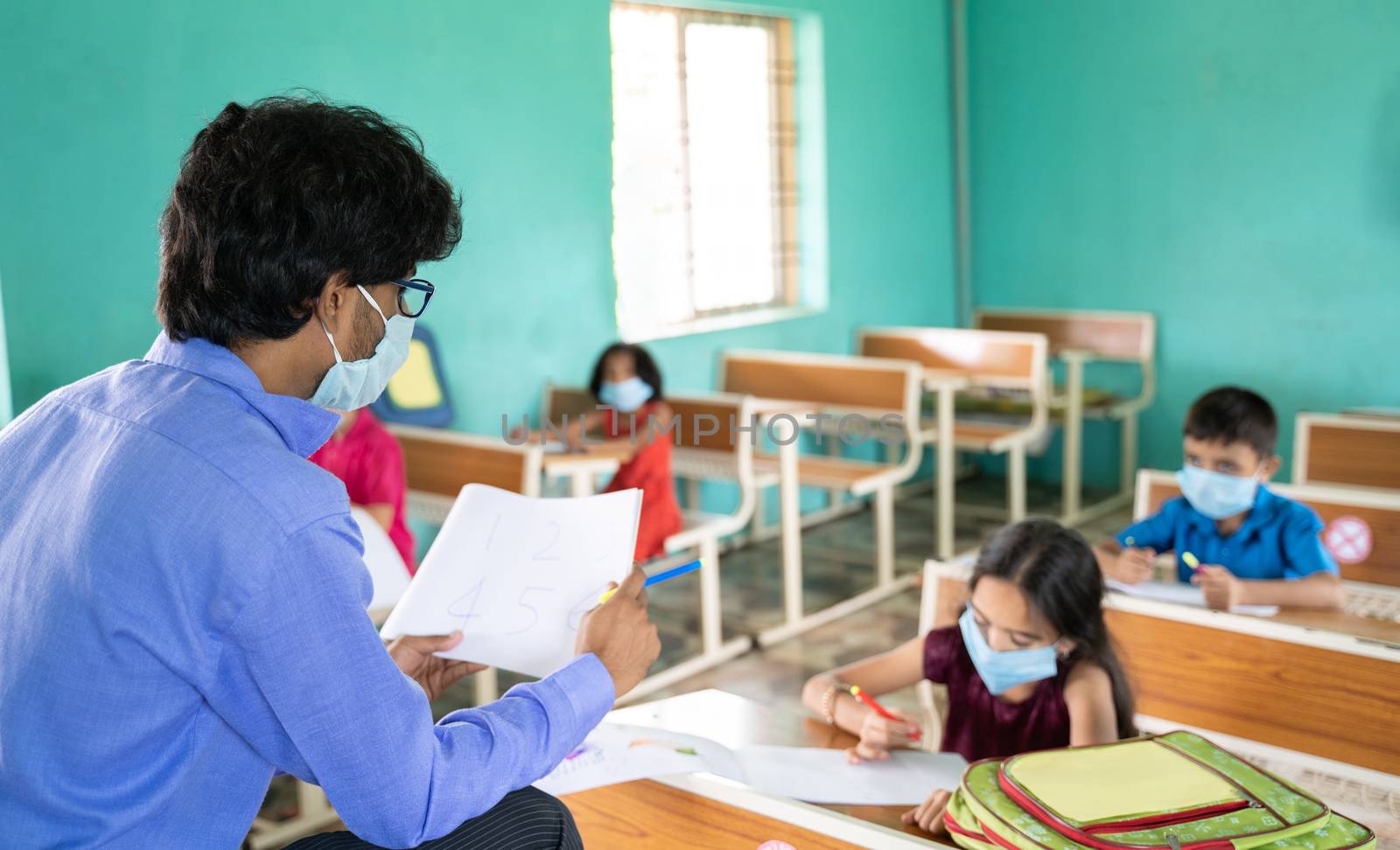 Teacher in medical mask with limited kids teaching at classroom while maintaining social distance due to coronavirus or covid-19 pandemic by lakshmiprasad.maski@gmai.com