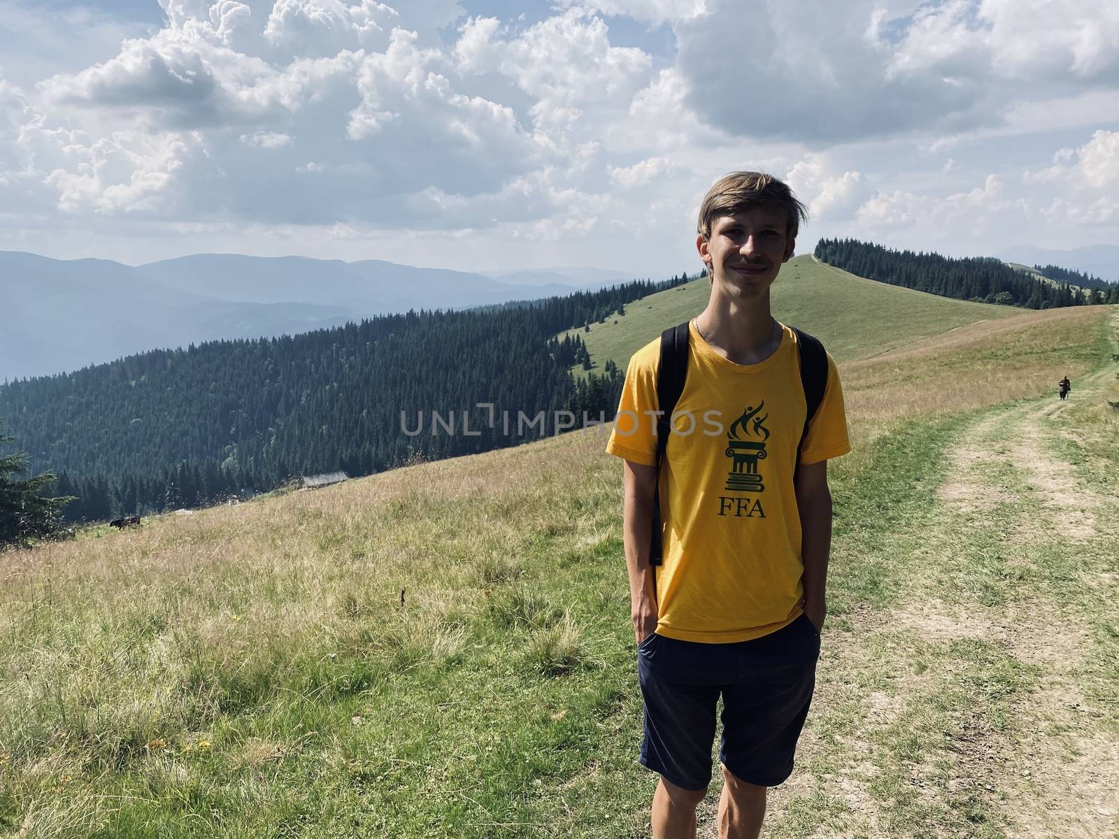 The young blonde guy is in the mountains. Blue sky and beautiful nature. Fit man climb. Green trees and green grass