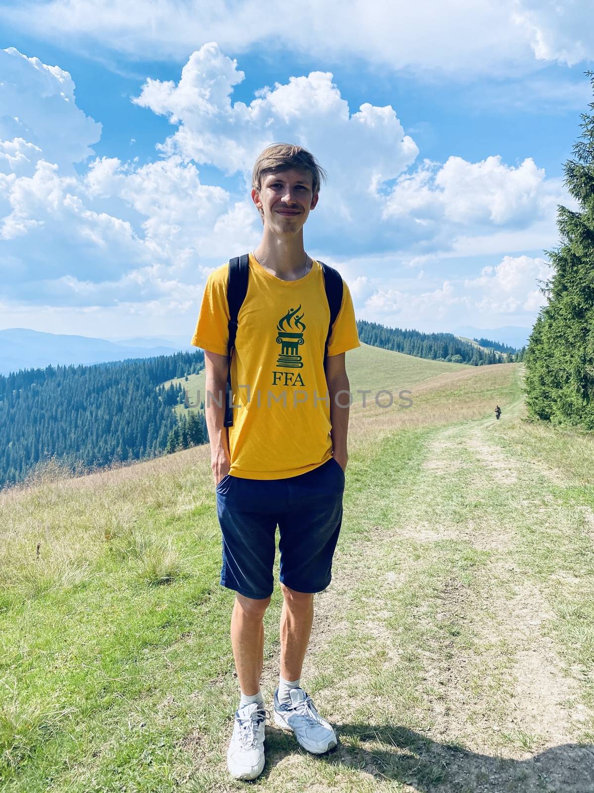 The young blonde guy is in the mountains. Blue sky and beautiful nature. Fit man climb. Green trees and green grass