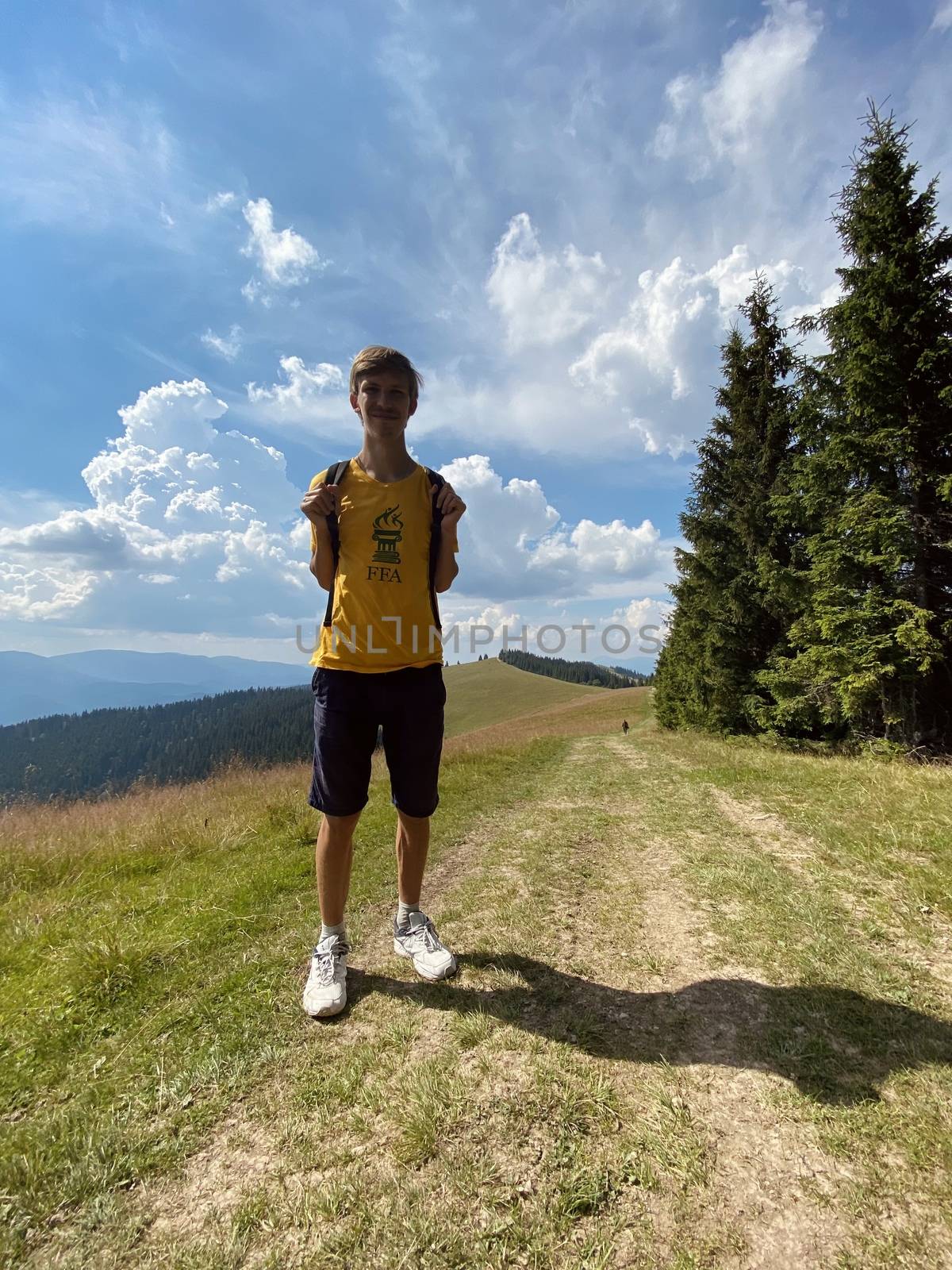 The young blonde guy is in the mountains. Blue sky and beautiful nature. Fit man climb. Green trees and green grass