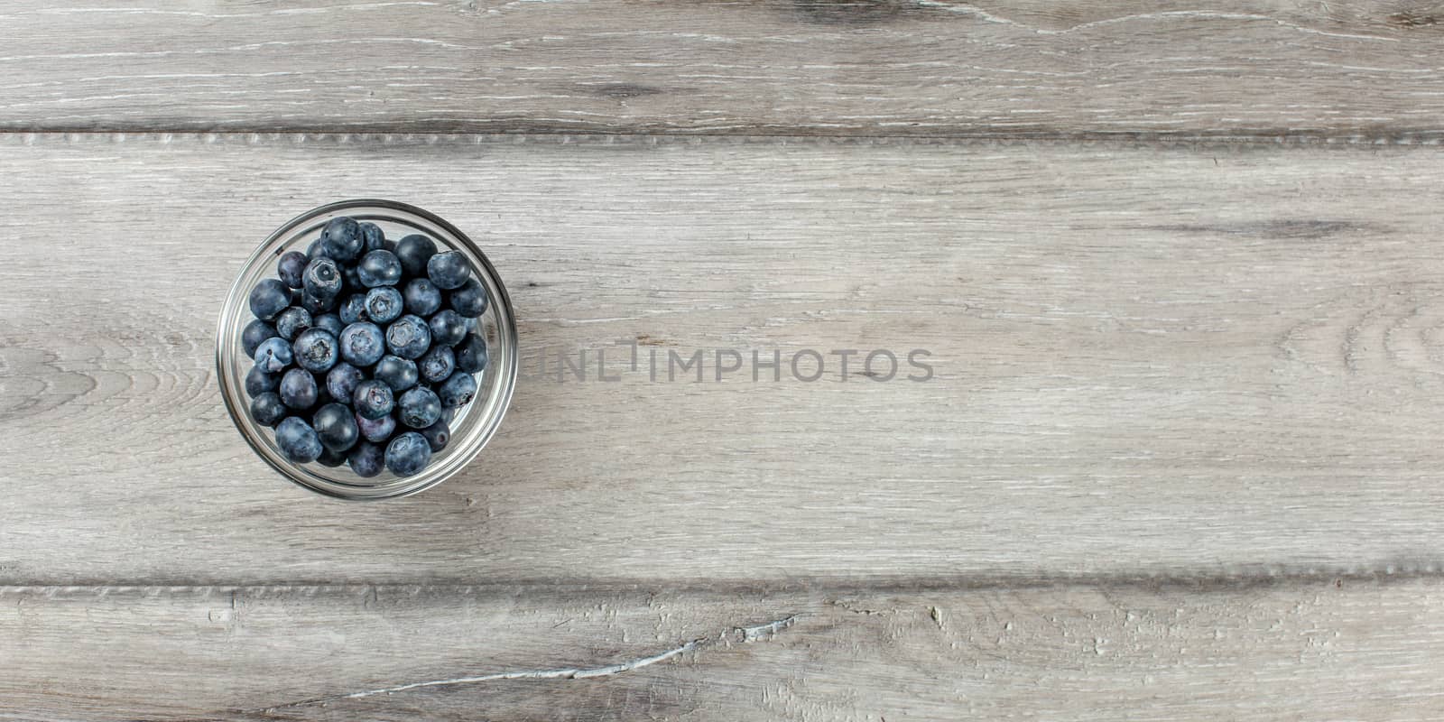 Wide shot from above to small glass cup full of blueberries on gray wood desk, with space for text.