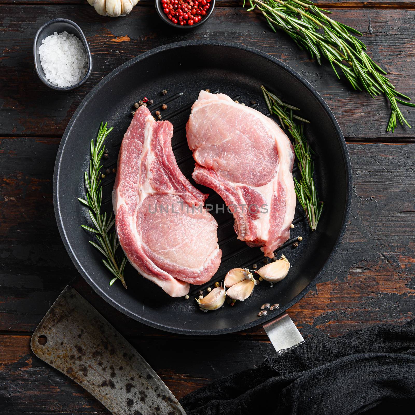 Raw pork steak on the bone in frying grill pan with butcher knife or cleaver and ingedients for grill top view square on vintage dark wood table.