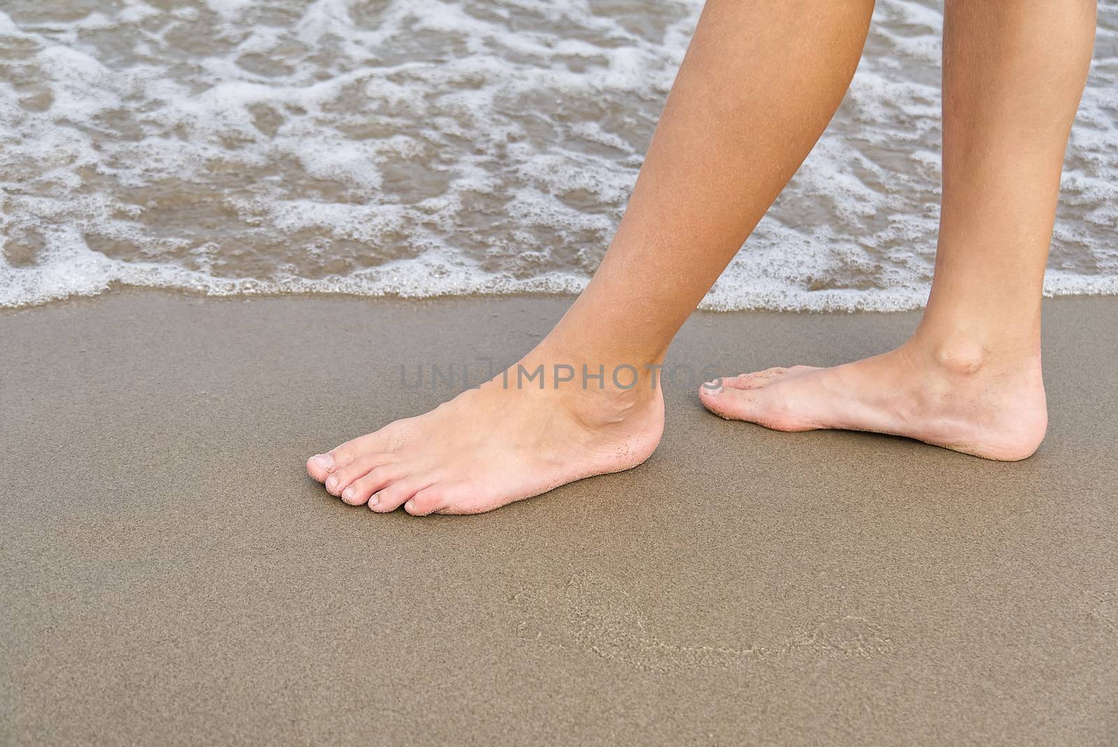 Close-up of child's legs walking on sand coast and sea water. Young girl legs on baltic sea beach. Teenager legs near sea line. by PhotoTime