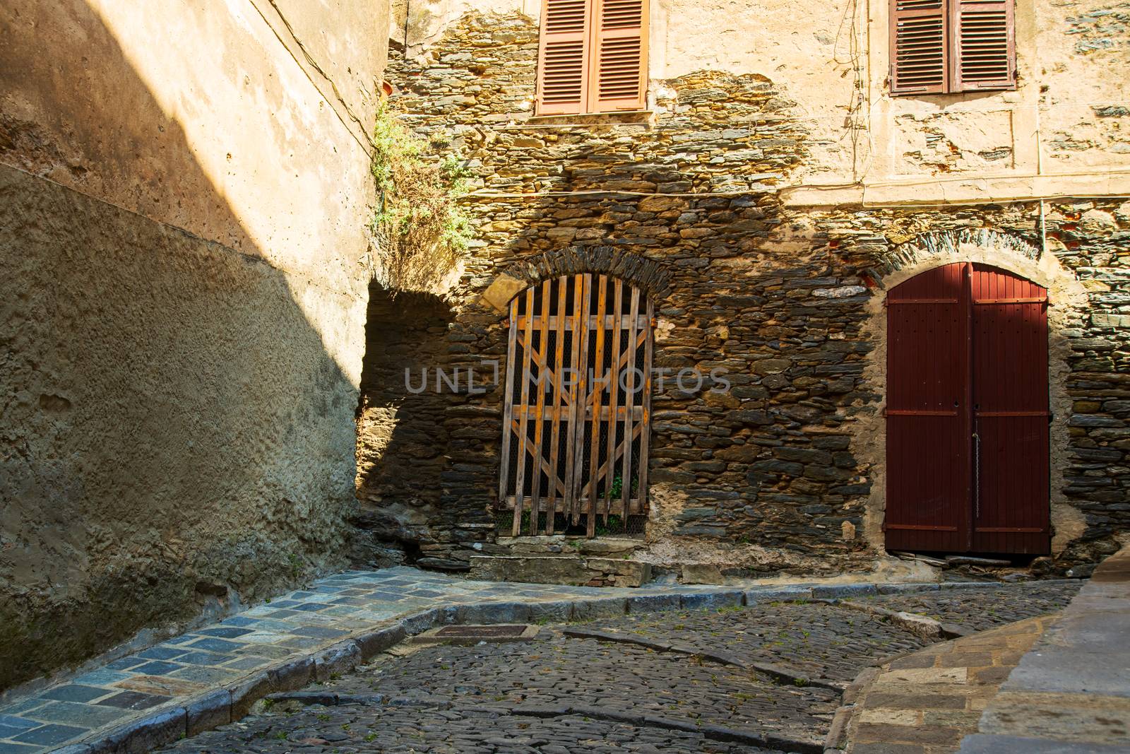 narrow alley with cobblestones in  a village on the isle of Corsica in France during summertime with a nice sunlight on the walls. The entrance of the houses are old wooden ports