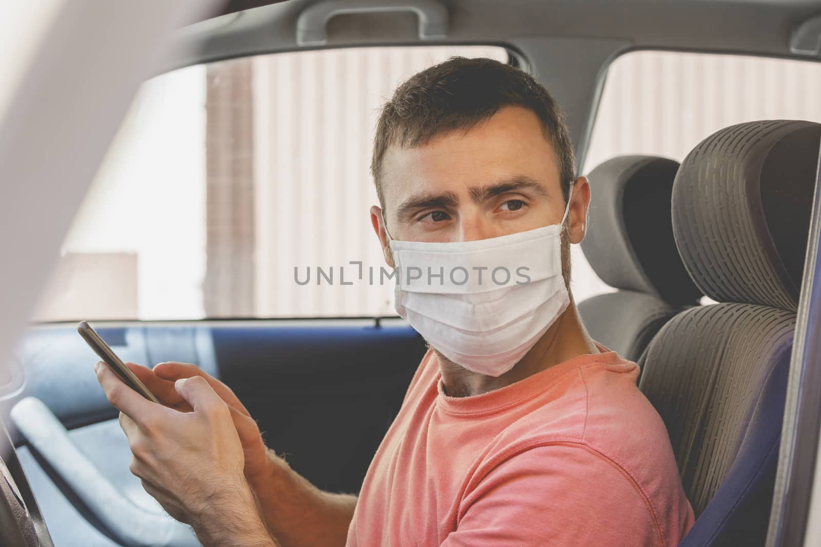 A young man, wearing a cloth face mask, checks the mobile phone inside the car by alvarobueno