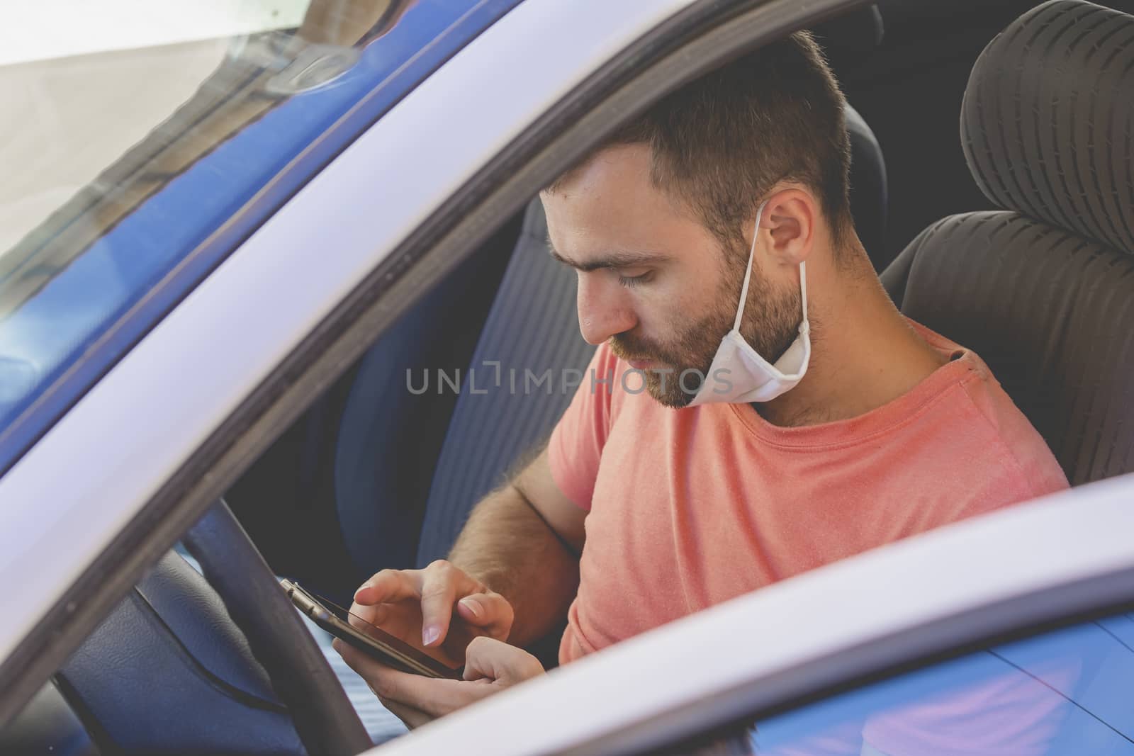 A young man, wearing a cloth face mask, checks the mobile phone inside the car, while he waits for someone before drive, in the province of Zaragoza, Spain.