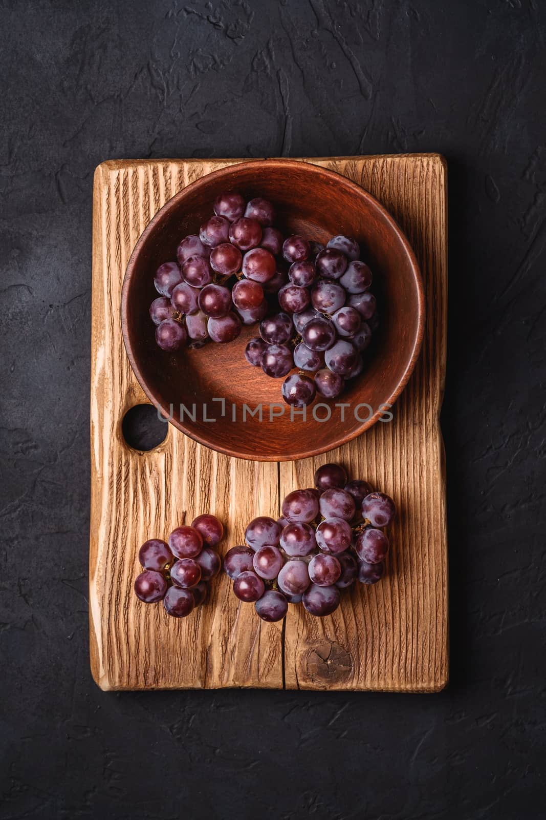 Fresh ripe grape berries in brown wooden bowl and cutting board on dark stone background, top view