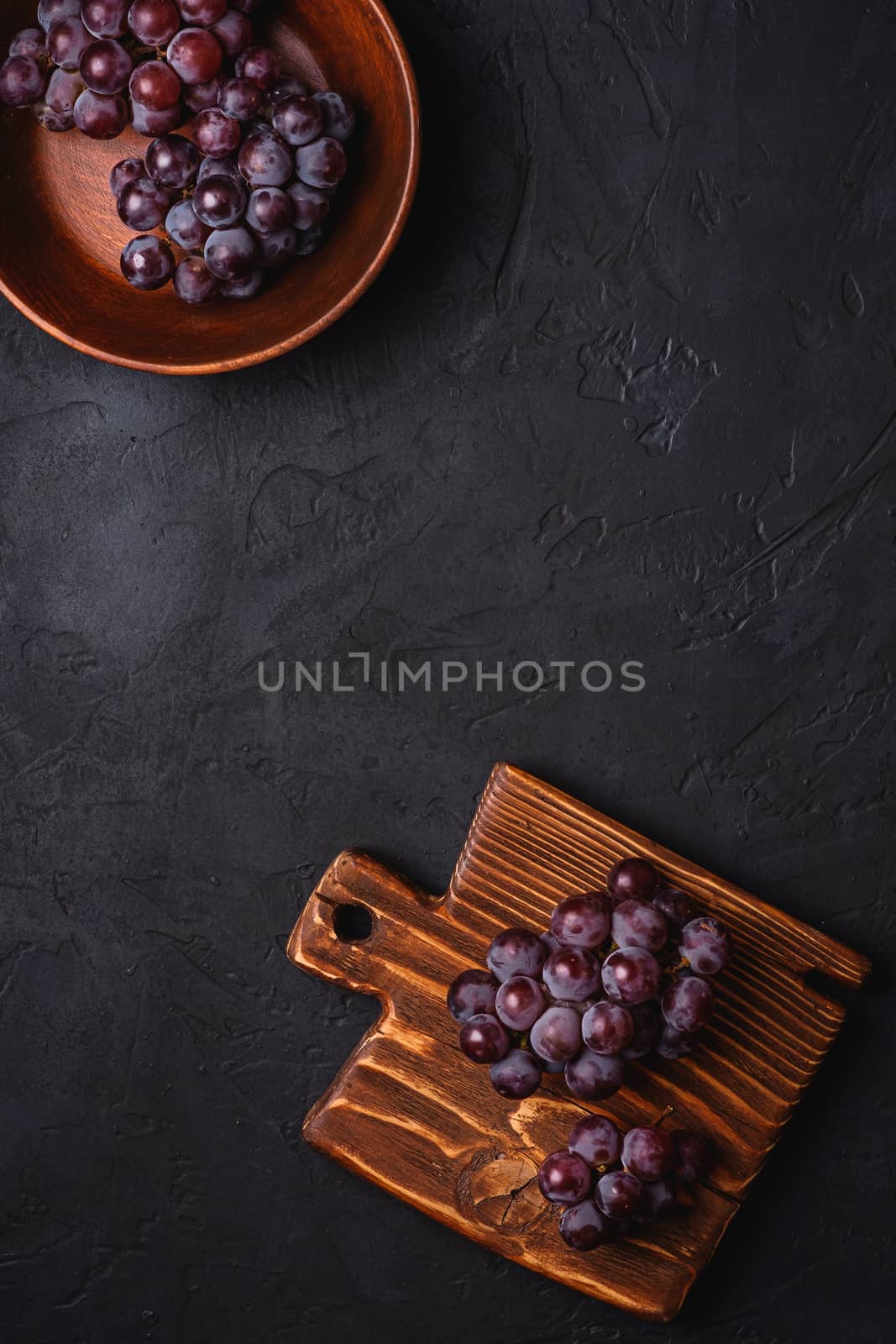 Fresh ripe grape berries in brown wooden bowl and cutting board on dark stone background, top view copy space