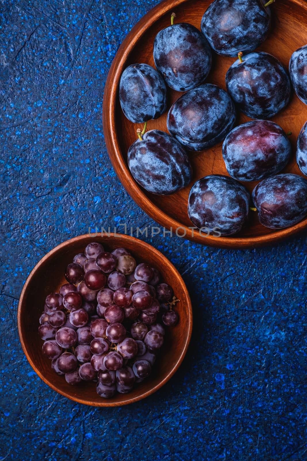 Fresh ripe plum fruits in brown wooden plate and grape berries in bowl on blue abstract background, top view