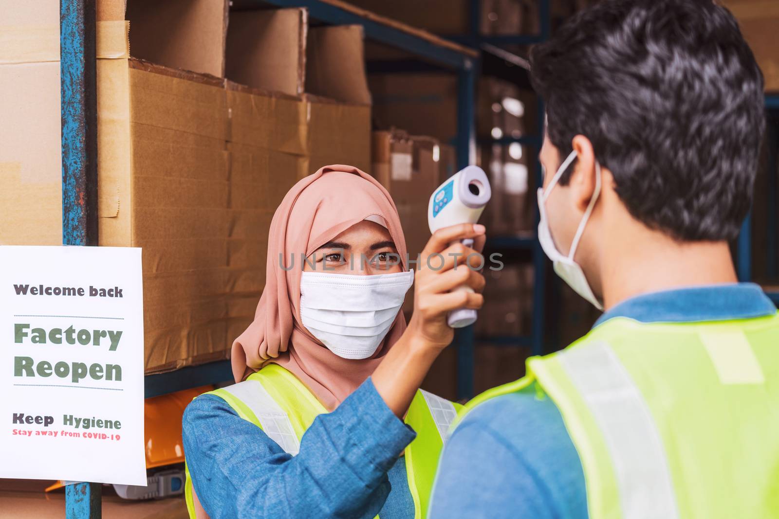 Muslim woman worker wearing surgical mask and Hijab uses Medical Digital Infrared Thermometer measure temperature to Indian man worker with safety clothes before start to work after warehouse reopen