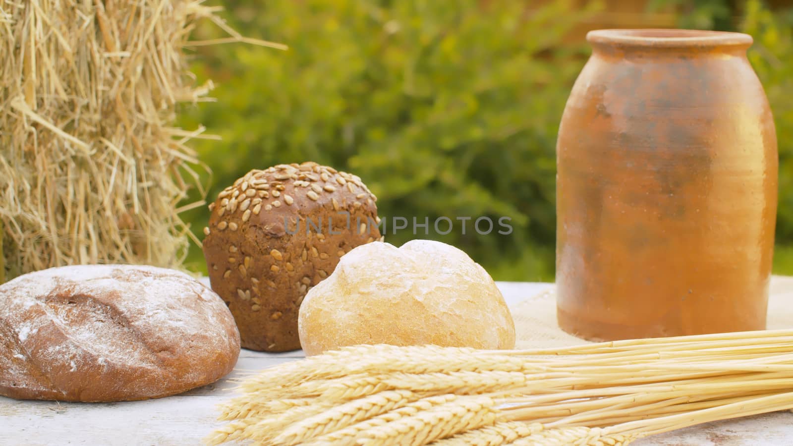 Close up loafs of traditional baked bread, ripe wheat ears, fresh hay and clay pot on table outdoors. Natural organic handmade food. Rural scene. Close-up shot with slide or move camera