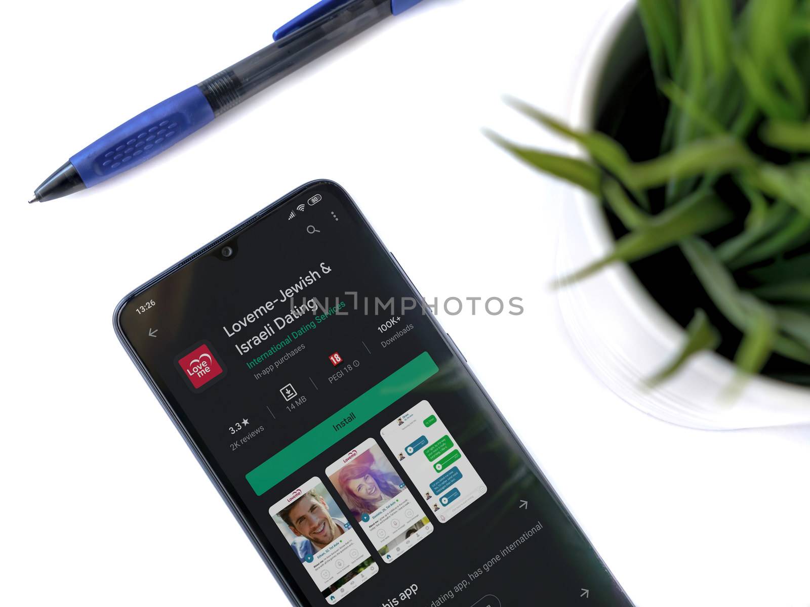 Lod, Israel - July 8, 2020: Modern minimalist office workspace with black mobile smartphone with Loveme app play store page on white background. Top view flat lay with copy space.