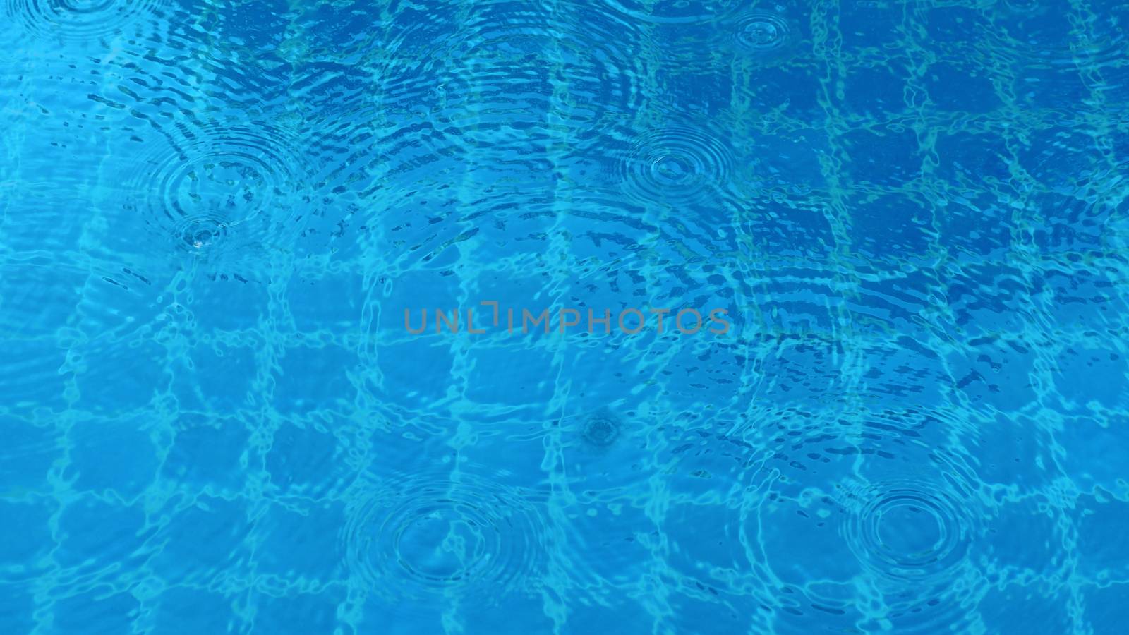 Rain drops on blue water swimming pool top view and sun light reflect on the texture.