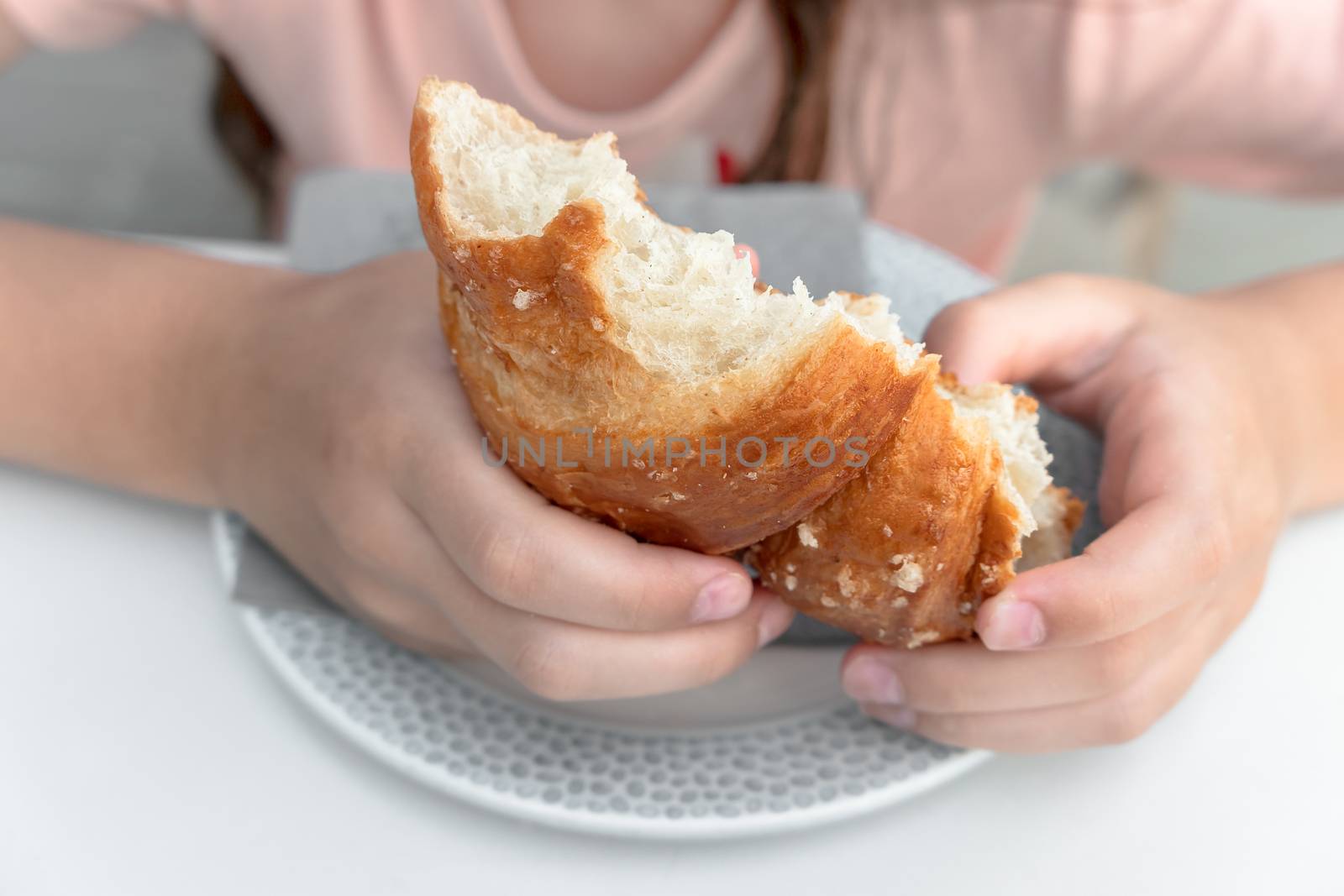 Pretty little girl eats a fresh croissant for breakfast. Portrait of cute girl with yummy face enjoy eating croissant. Healthy food for children concept.