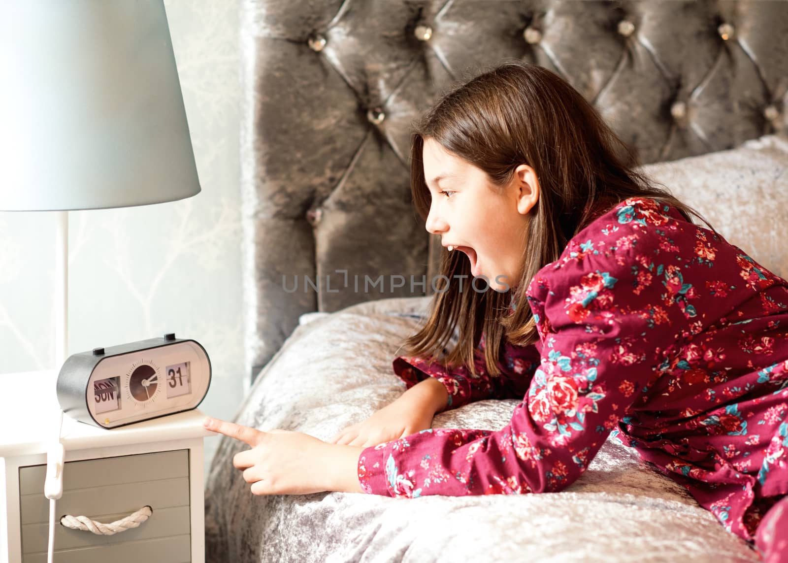 A teenager in pajamas lying in bed and looking at a clock. She is late for school.