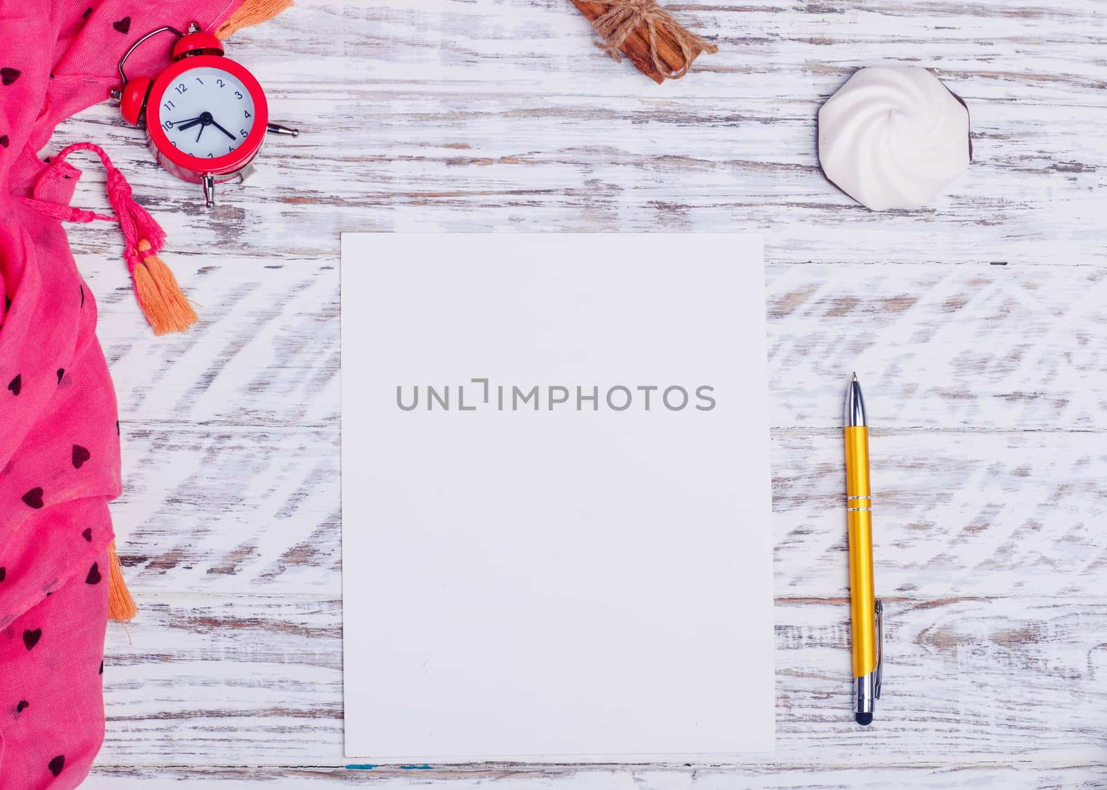 Mockup of white empty paper, yellow pen, clock, sweets, and pink scarf on wooden desk. Flat Lay with no people. View from the top.