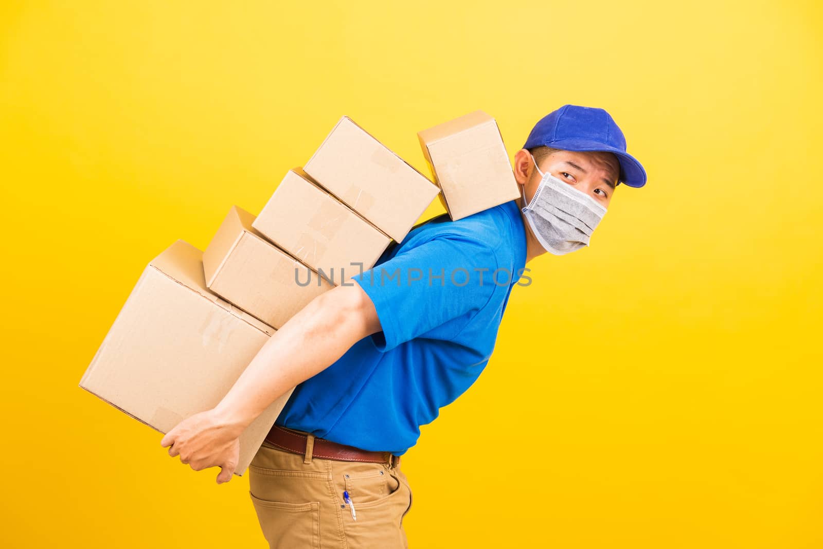 Asian young delivery worker man in uniform wearing face mask protective he has many job lifting stack heavy a lot of boxes on back, under coronavirus COVID-19, studio shot isolated yellow background