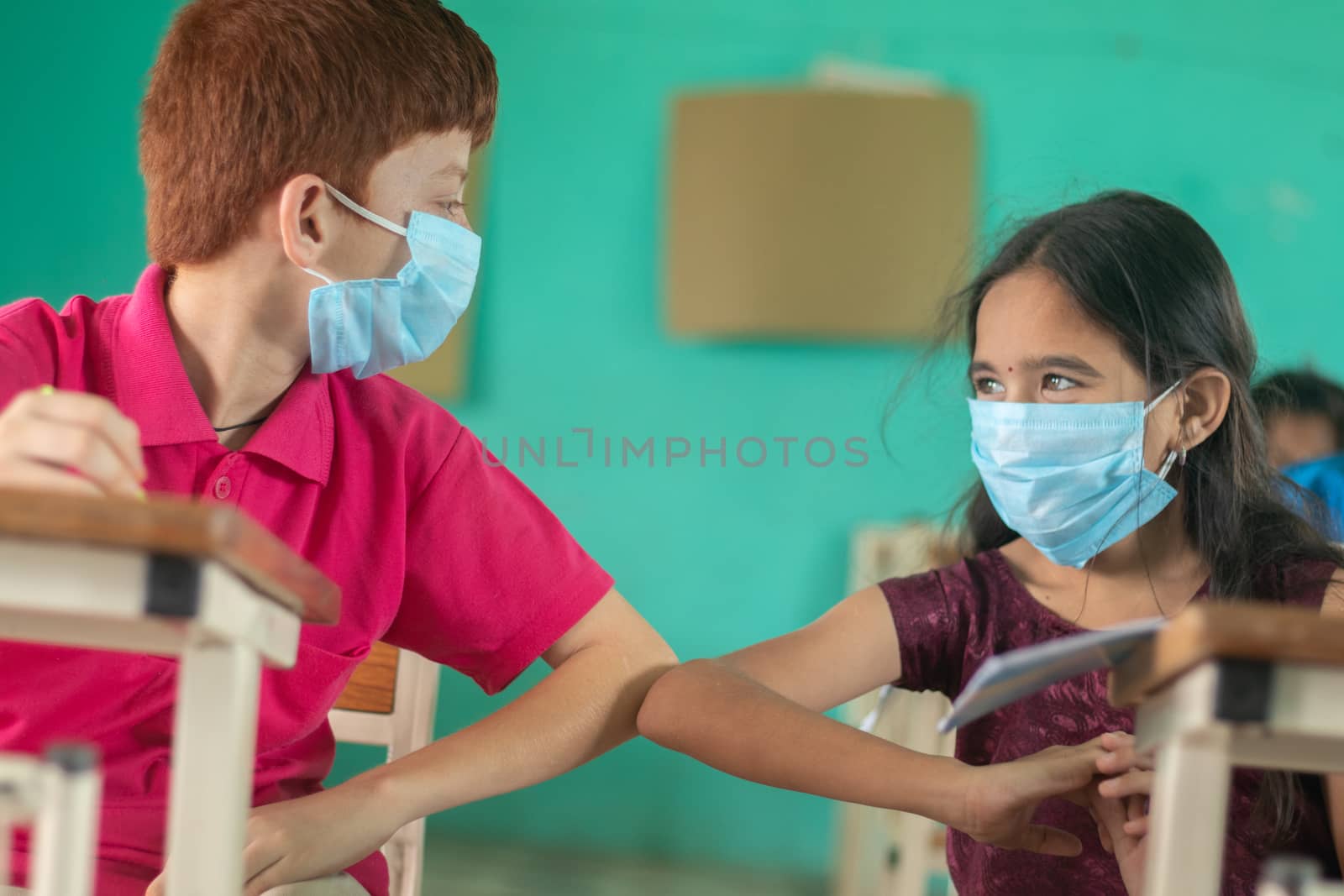 Two kid in medical mask at classroom greeting each other with elbow bumps while maintaining socail distance at school - Concept of school reopen, back to school safety measures and new normal lifestye by lakshmiprasad.maski@gmai.com