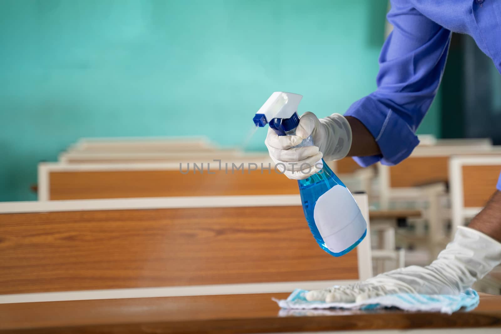 Close up of hands with gloves disinfecting desk by using sanitizer at classroom - Cleaning dust on table surface with cloth and Disinfectant Spray to protect from coronavirus or covid-19 infection. by lakshmiprasad.maski@gmai.com