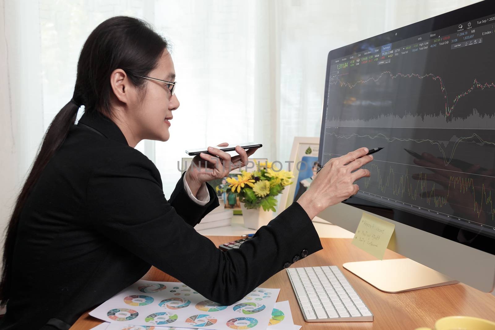 Business women holding smart phone to speak with partner by speaker phone and analysis stock chart on computer PC screen at office. Business people call for working with customer or partner with smart phone. Business concept.