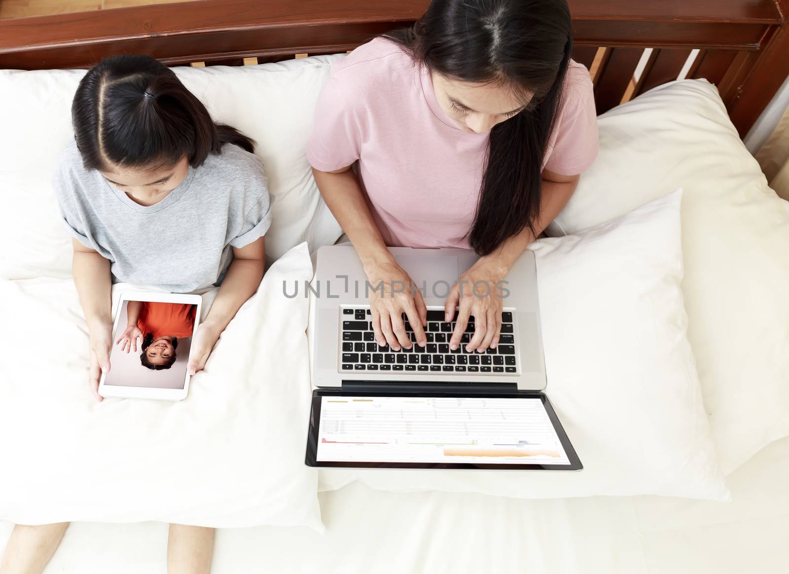 Top view Asian mother and daugther looking laptop and tablet on bed in bedroom. Business women woking with laptop at home with her daughter enjoy speak with friend video call  on bed. soft focus photo
