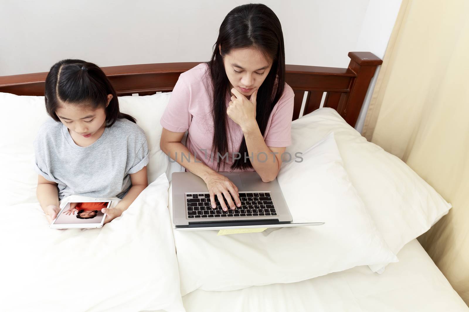 Top view Asian mother and daugther looking laptop and tablet on bed in bedroom. Business women woking with laptop at home with her daughter enjoy speak with friend video call  on bed. soft focus photo