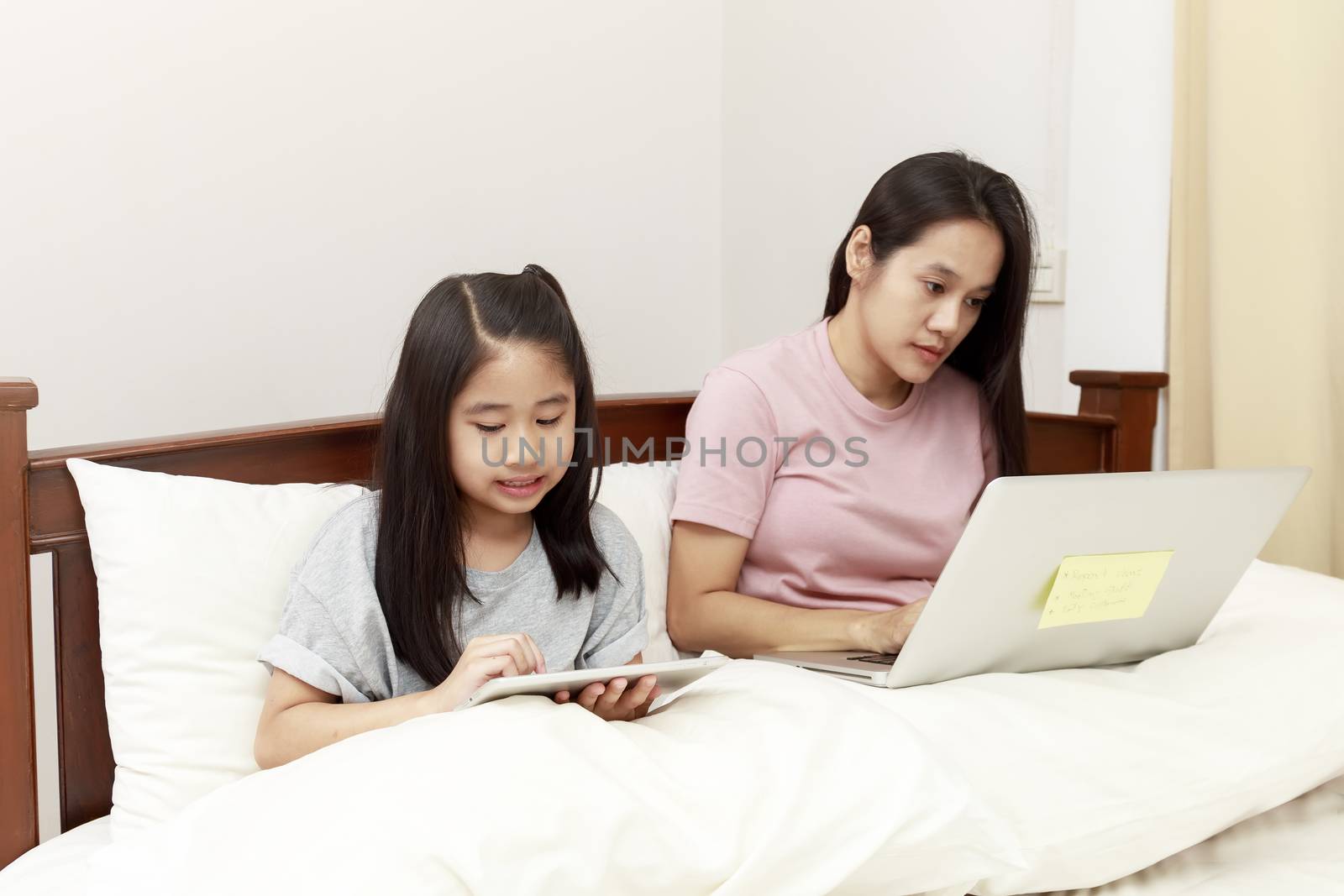 Asian mother and daugther looking laptop and tablet on bed in bedroom. Business women woking with laptop at home with her daugther smiling enjoy with tablet on bed. Work at home concept.