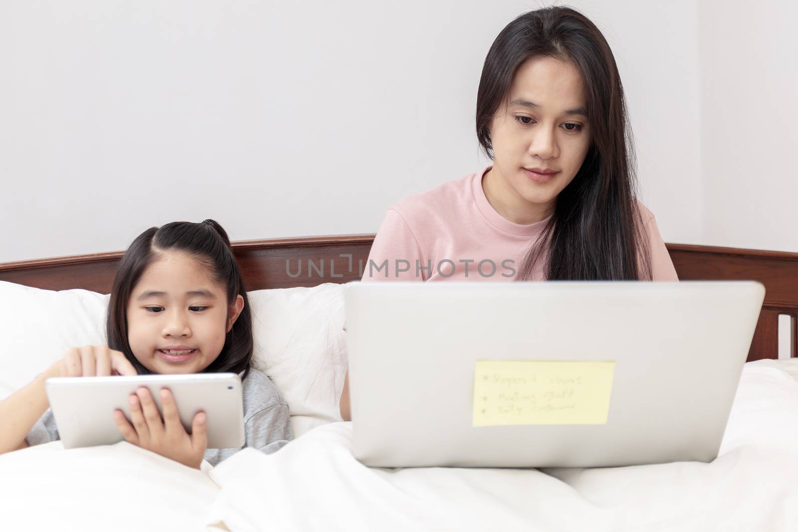 Asian mother and daugther looking laptop and tablet on bed in bedroom. Business women woking with laptop at home with her daugther smiling enjoy with tablet on bed. Work at home concept.