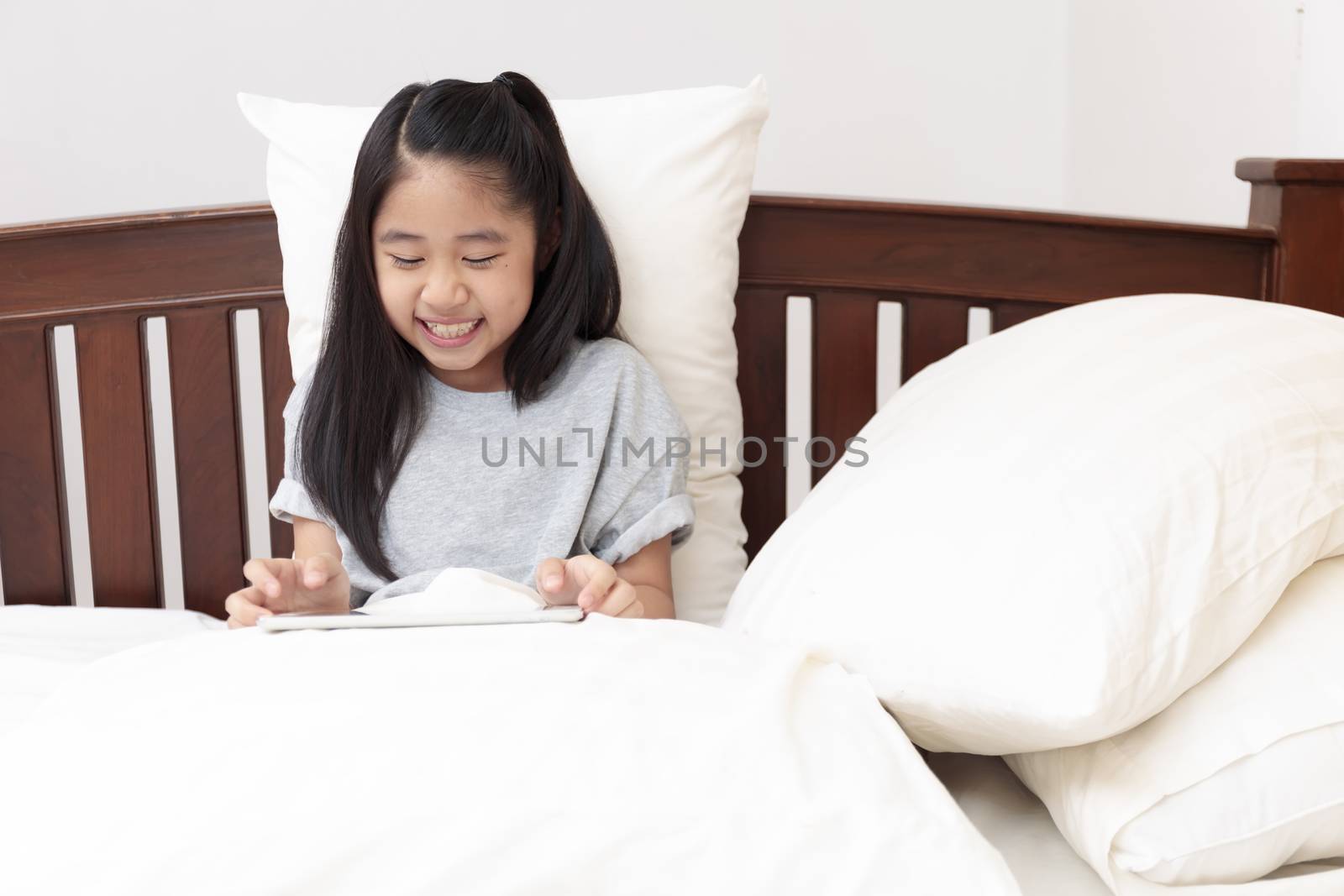Asian little girl smiling in hands holding and play tablet on bed in bedroom. Little girl enjoy with tablet on bed. Asian girl looking and play tablet.