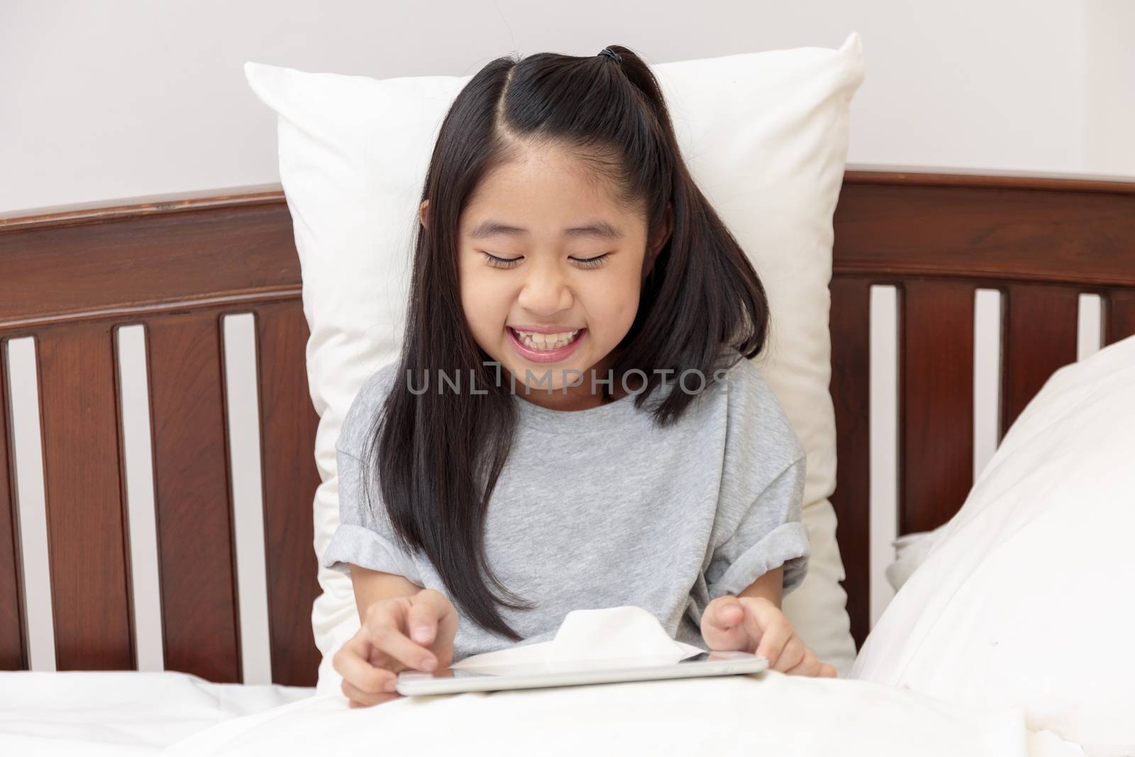 Asian little girl smiling in hands holding and play tablet on bed in bedroom. Little girl enjoy with tablet on bed. Asian girl looking and play tablet.