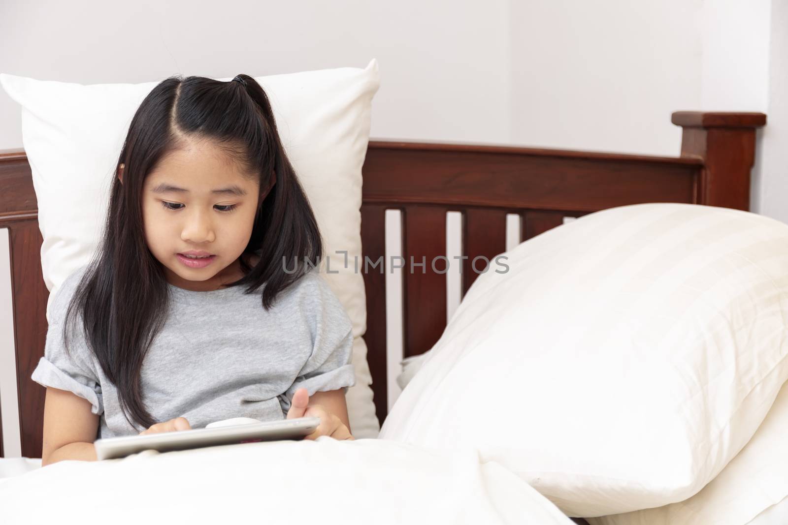 Asian little girl smiling in hands holding and play tablet on bed in bedroom. Little girl enjoy with tablet on bed. Asian girl looking and play tablet.