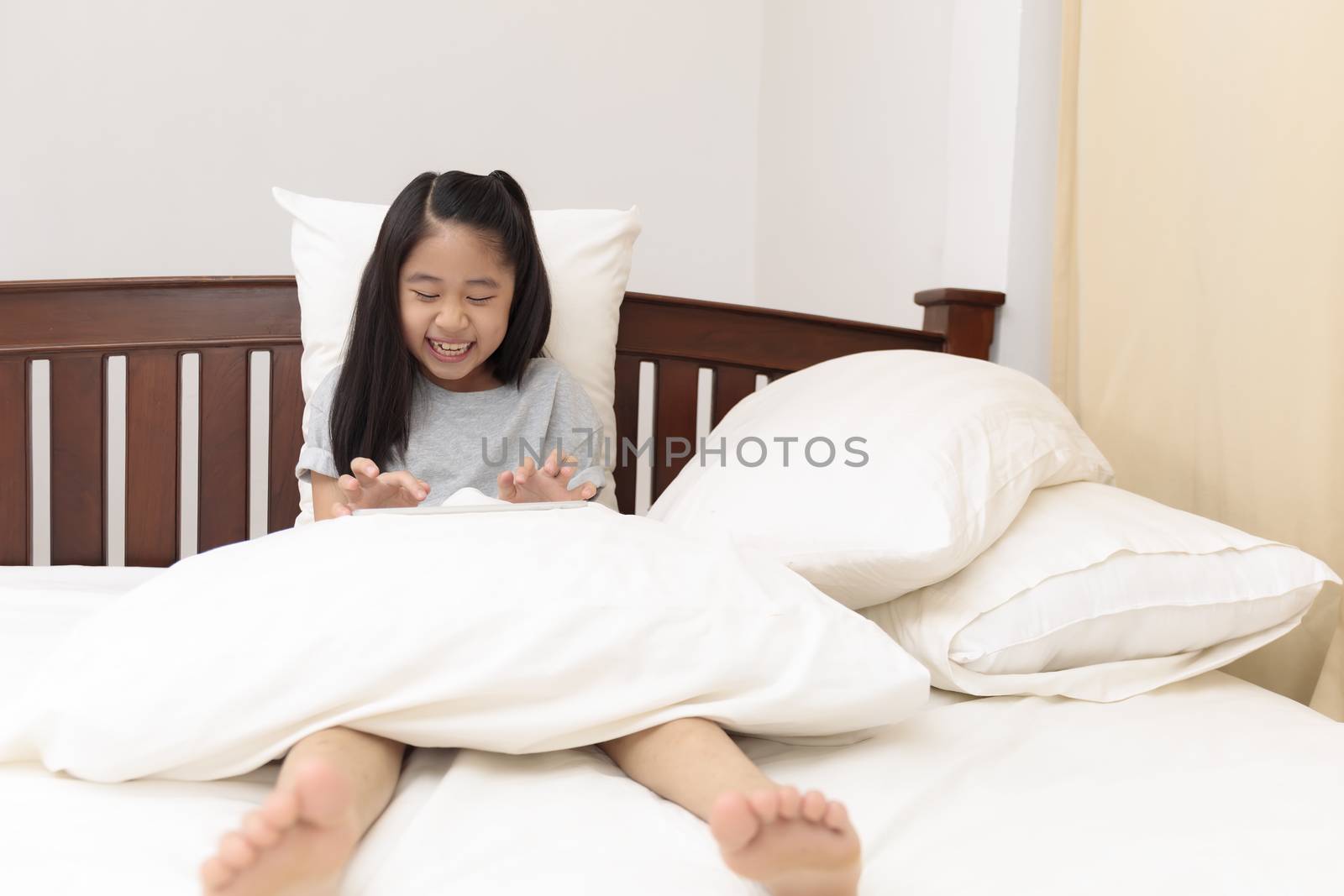 Asian little girl smiling in hands holding and play tablet on bed in bedroom. Little girl enjoy and funny with tablet on bed. Asian girl looking and play tablet.