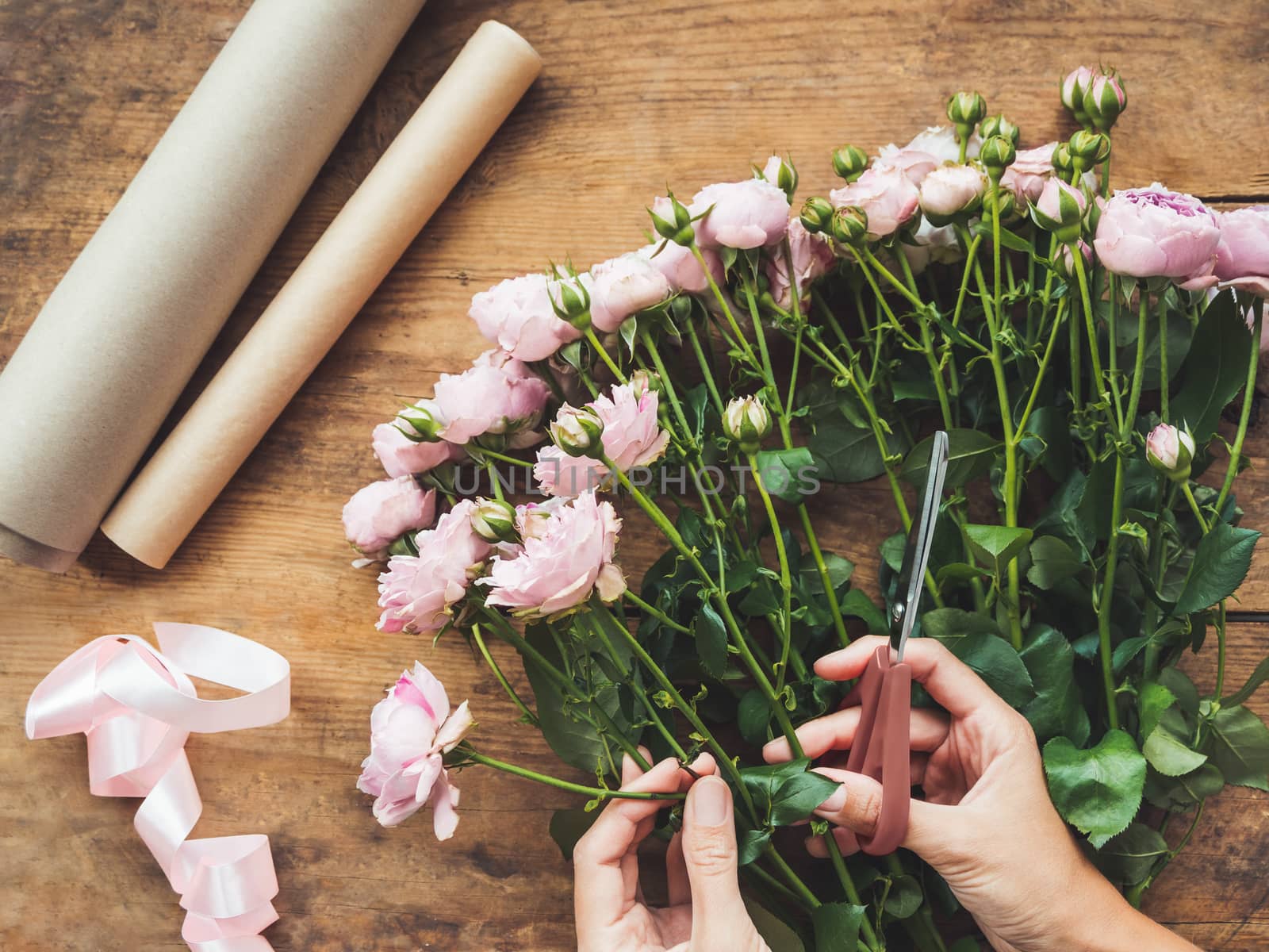 Top view on wooden table with roses, scissors, craft paper and pink ribbon on it. Florist work place. Accessories for making bouquets and floral compositions.