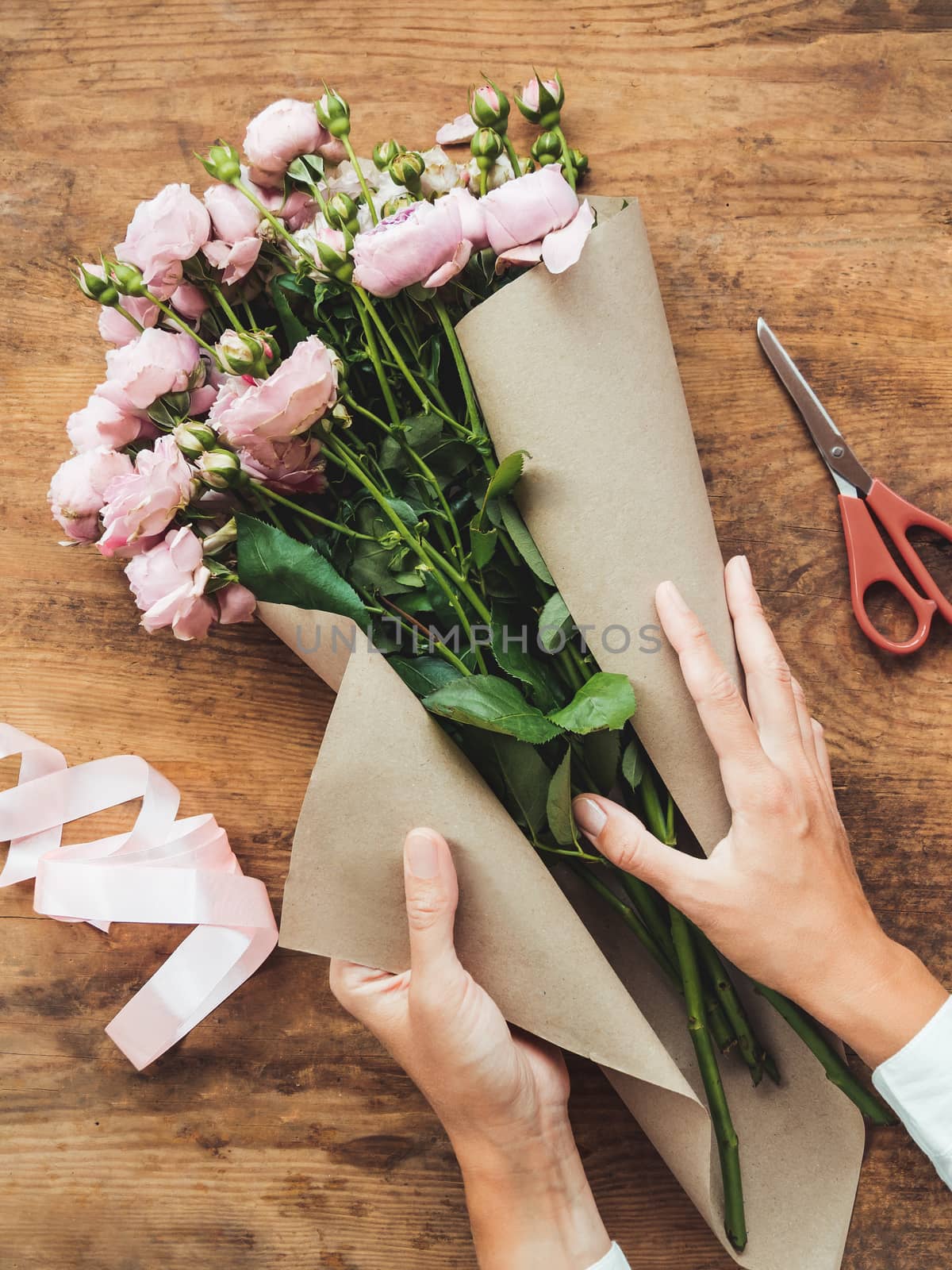 Top view on wooden table with roses, scissors, craft paper and pink ribbon on it. Florist work place. Accessories for making bouquets and floral compositions.