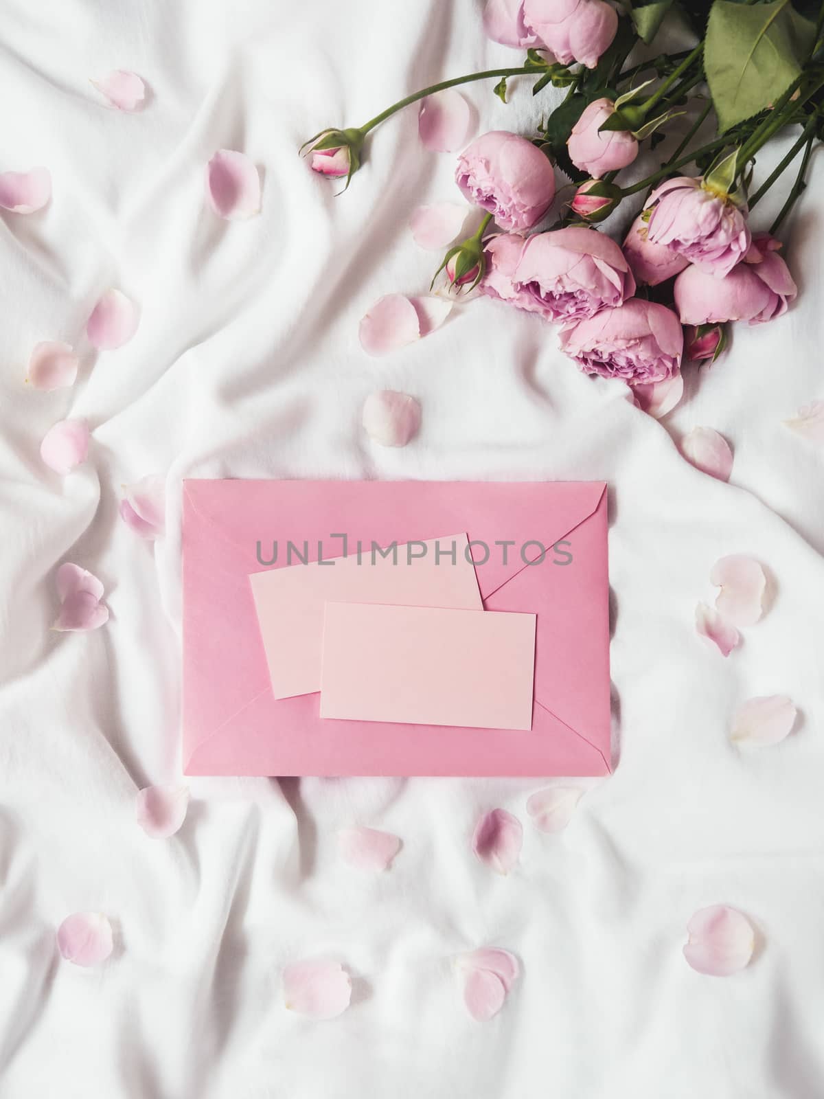 Roses and petals on crumpled white fabric. Natural elegant decoration. Romantic background with copy space on pink envelope and visit cards. Top view, flat lay.