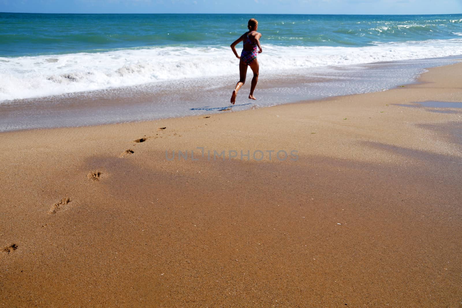 teenage girl runs along the seashore, leaving footprints on the wet sand, rear view