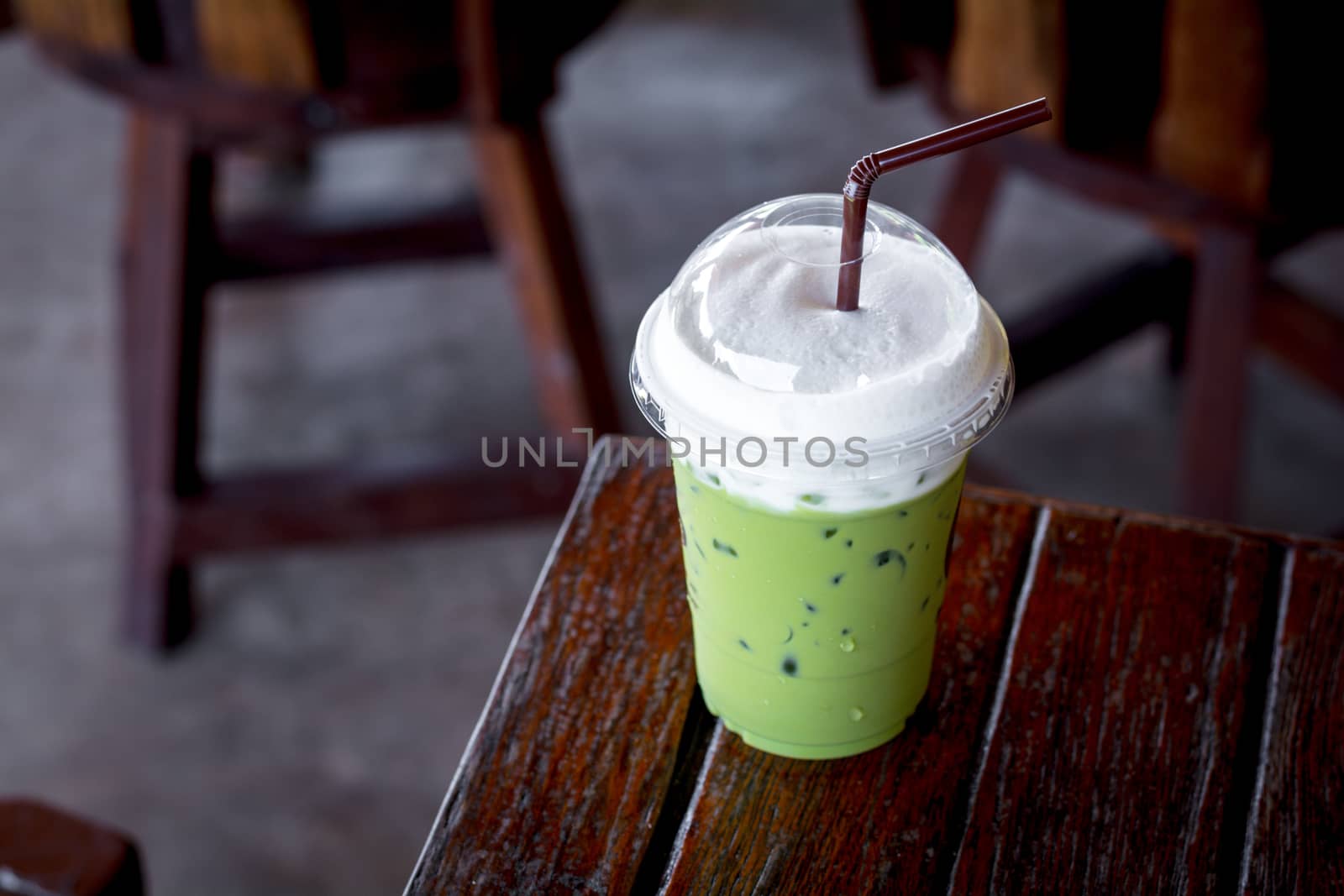 Iced green tea or matcha latte with milk foam in a plastic cup with brown straw on a wooden table in the coffee shop. Healthy drink and beverage concept.