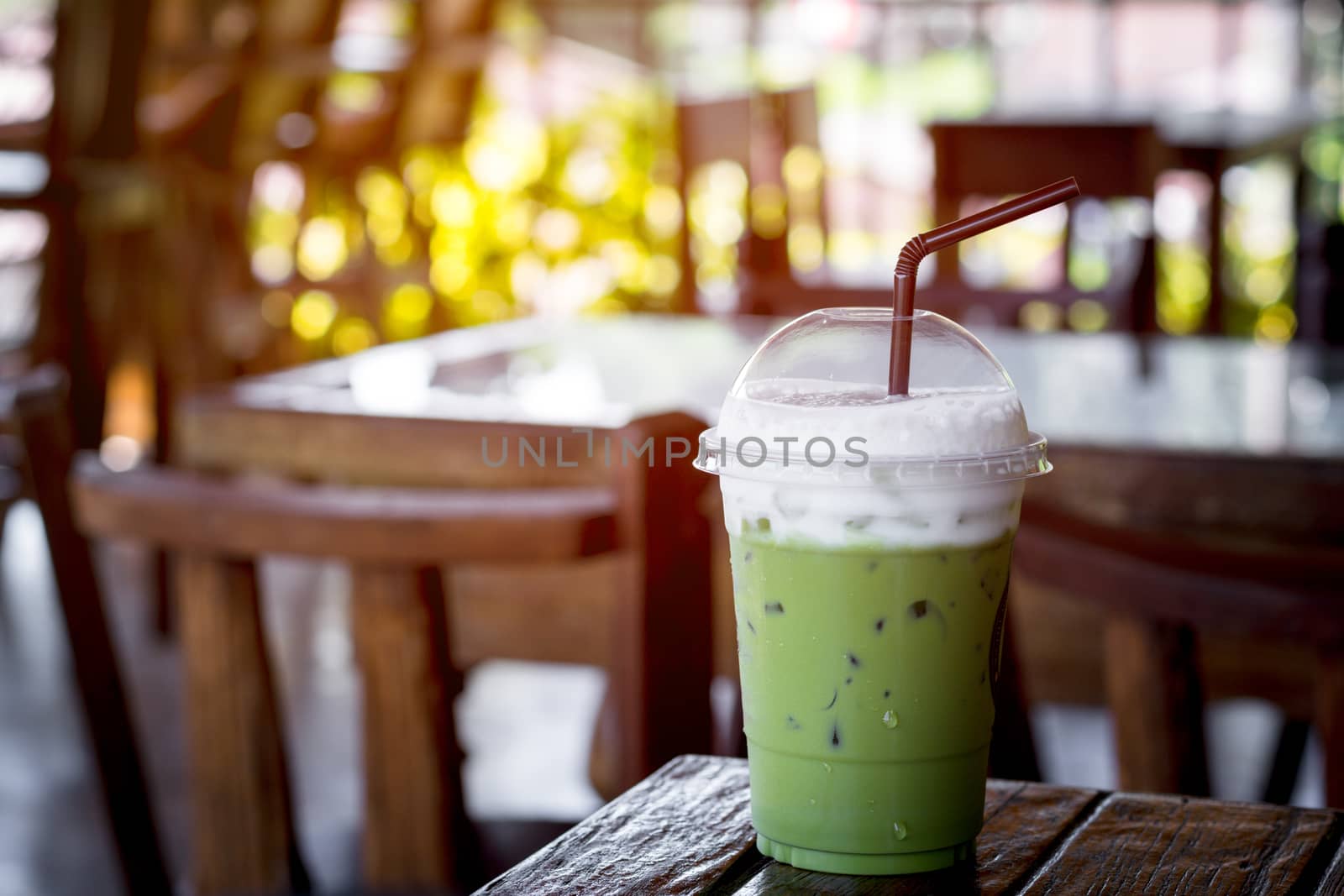 Iced green tea or matcha latte with milk foam in a plastic cup with brown straw on a wooden table in the coffee shop with sun light. Healthy drink and beverage concept.